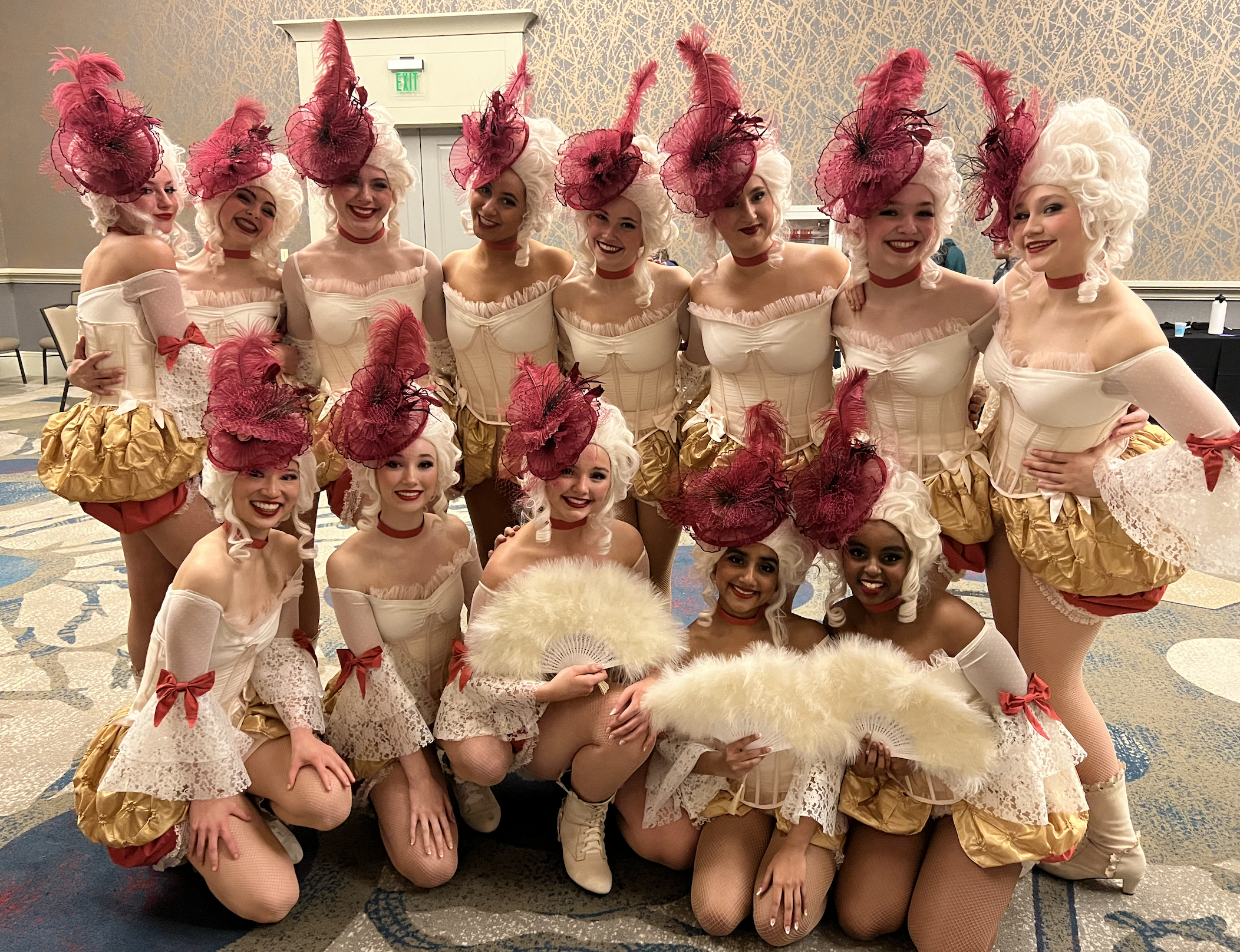 Group of women dressed in elaborate corset and skirt costumes, wearing large white wigs and red feathered hats at an indoor event.