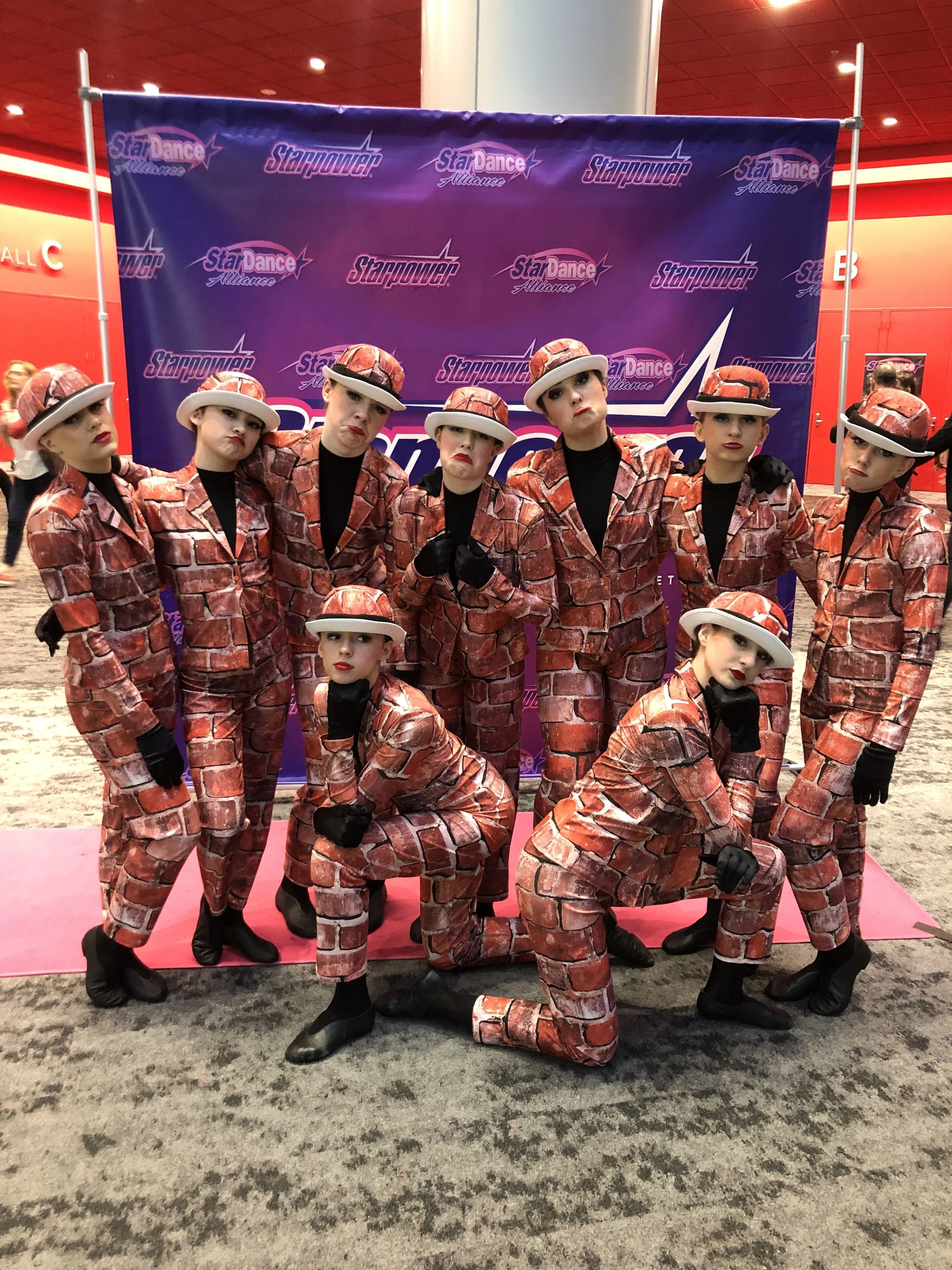 A group of seven kids dressed in matching dance costumes resembling bacon strips, posing in front of a purple backdrop with the Starpower Alliance logo. They are wearing matching bacon-themed jackets, hats, and gloves, with some in a crouched stance 
