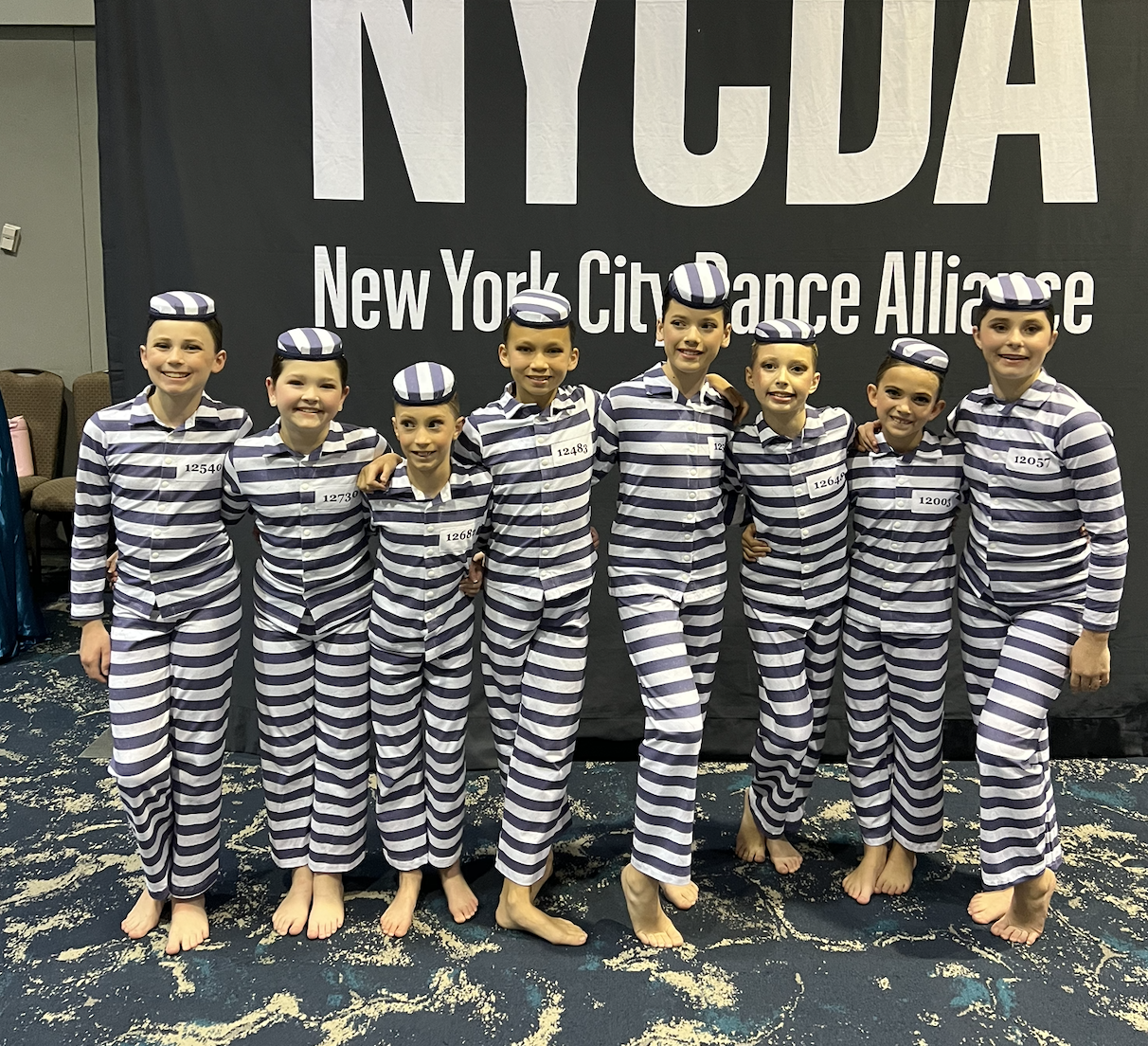 A group of young dancers in matching striped costumes and hats, standing arm-in-arm in front of a backdrop that reads 'NYCDA New York City Dance Alliance'.