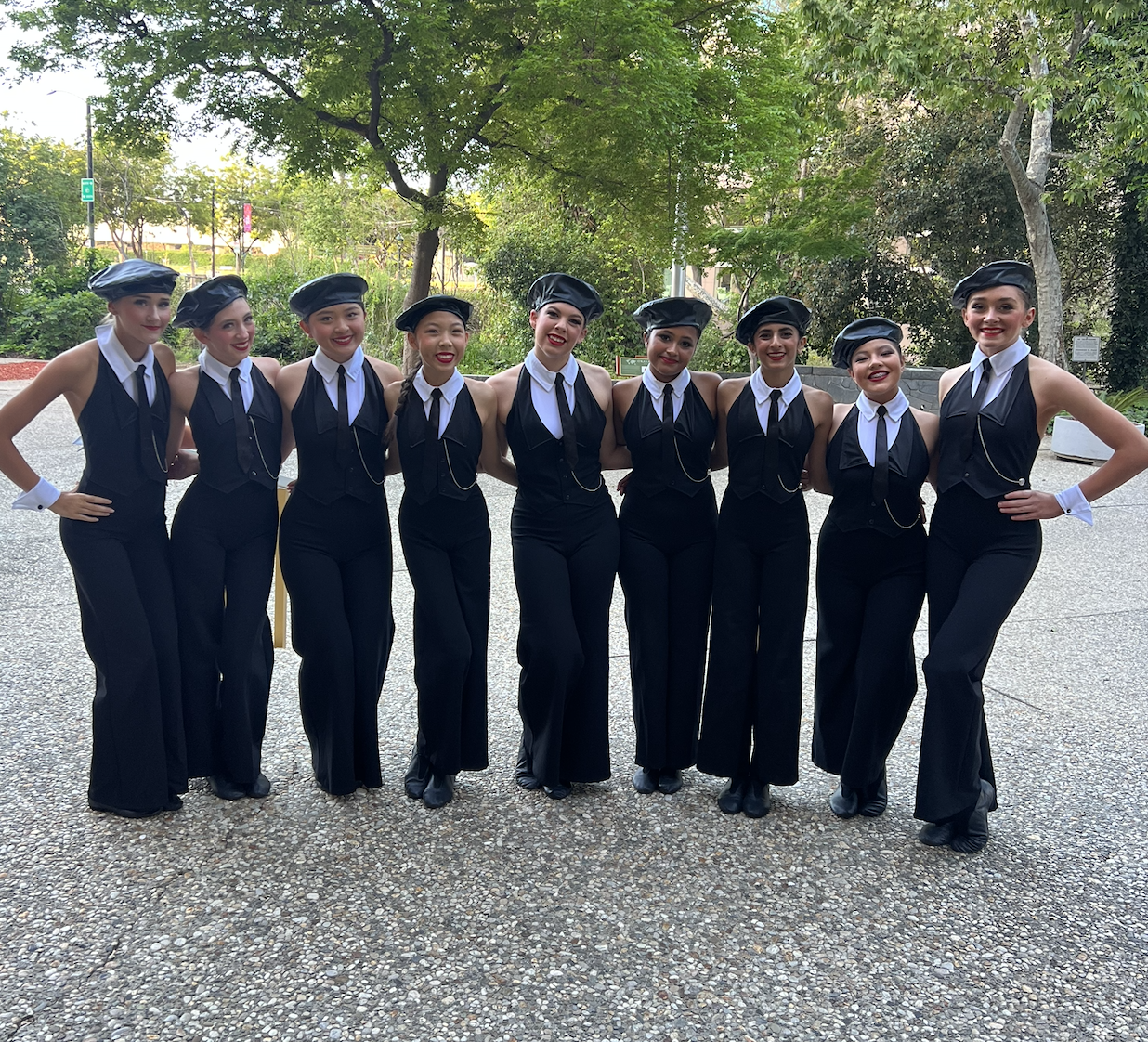 A group of nine women dressed in black pants, white shirts, black vests, black ties, and black berets posing outdoors with arms around each other's shoulders.