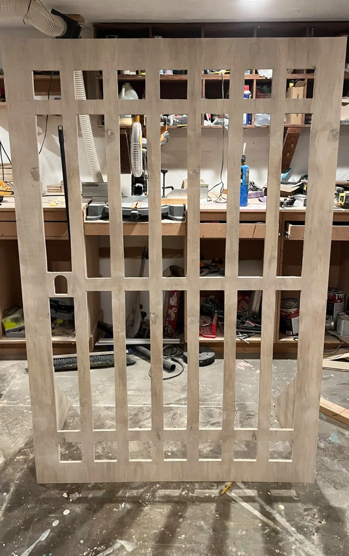 A wooden panel with rectangular cutouts, standing upright in a woodworking workshop, with tools and supplies visible in the background.