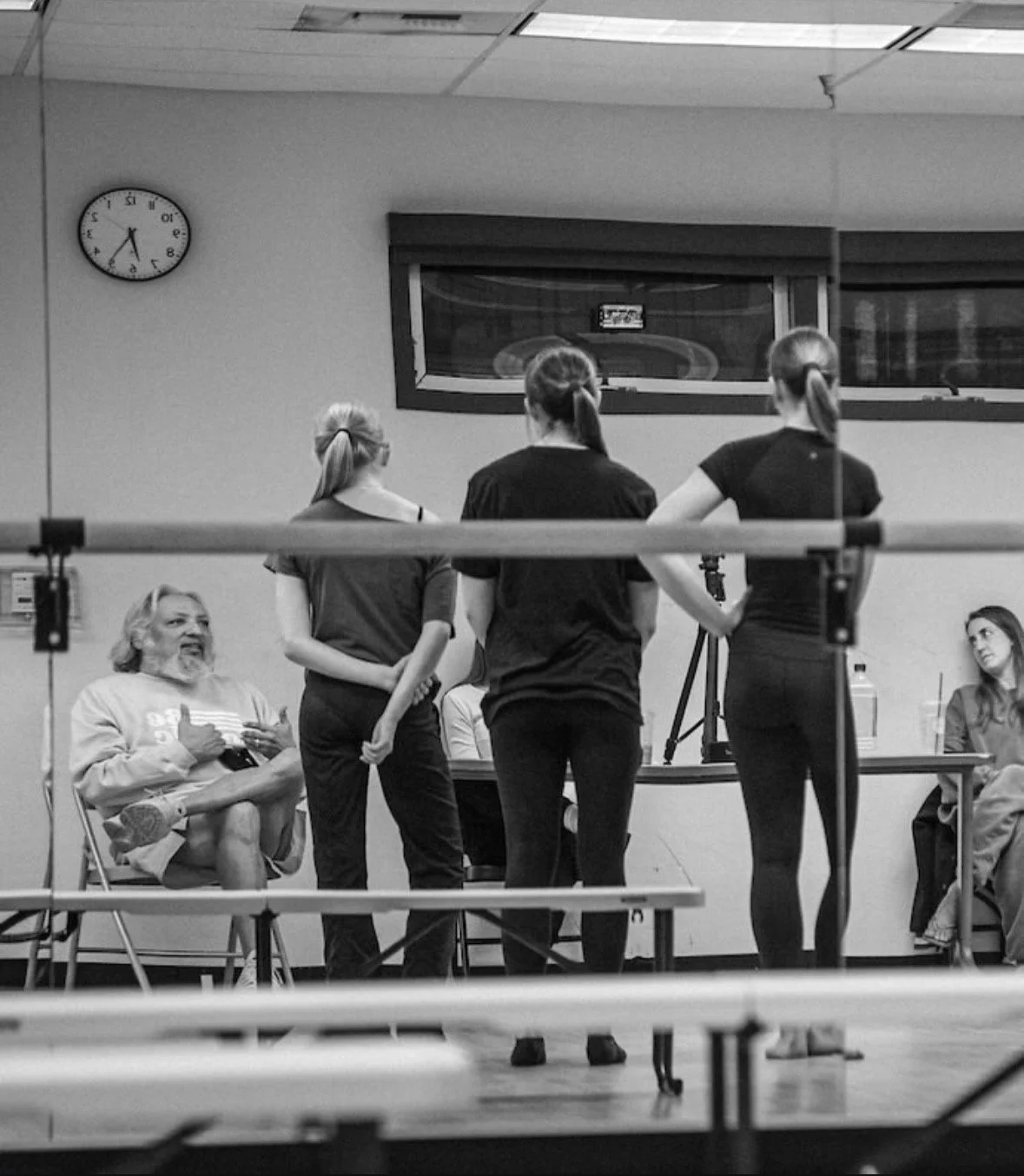 Three young women are standing in front of a seated man inside a room, possibly a gym or dance studio, with a clock showing 7:25 on the wall. The women are facing away from the camera, while the man is talking and smiling at them.