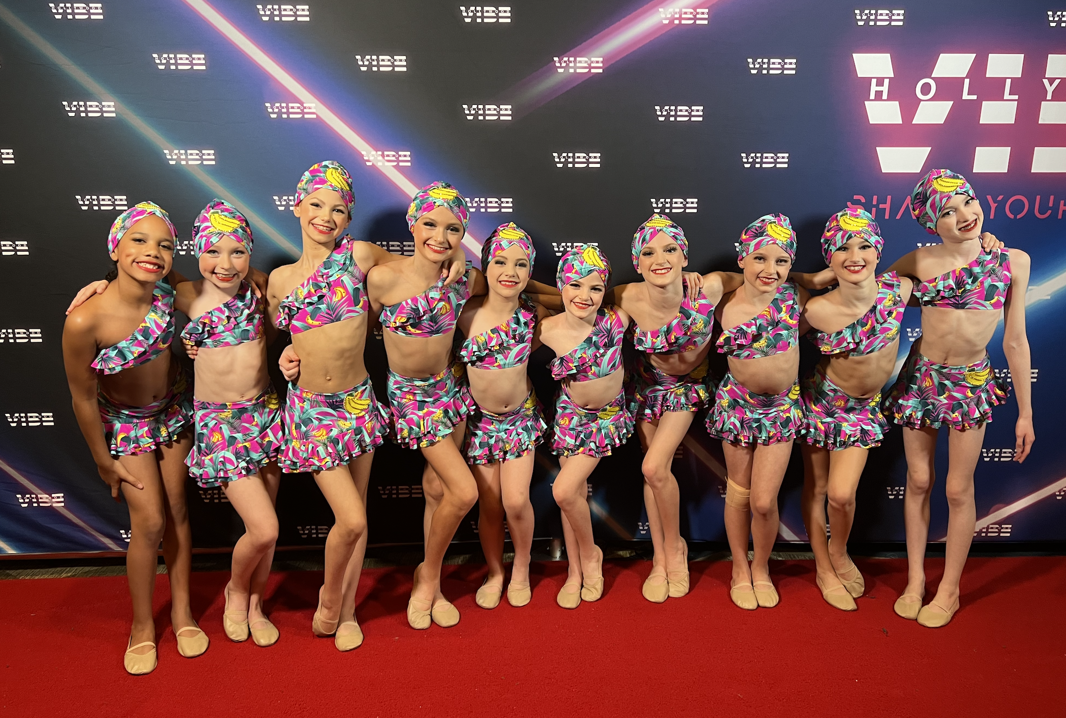 Group of young women in colorful matching costumes and headscarves, standing on a red carpet in front of a backdrop with logos for VIBE and HOLLYWOOD SHOW YOU.