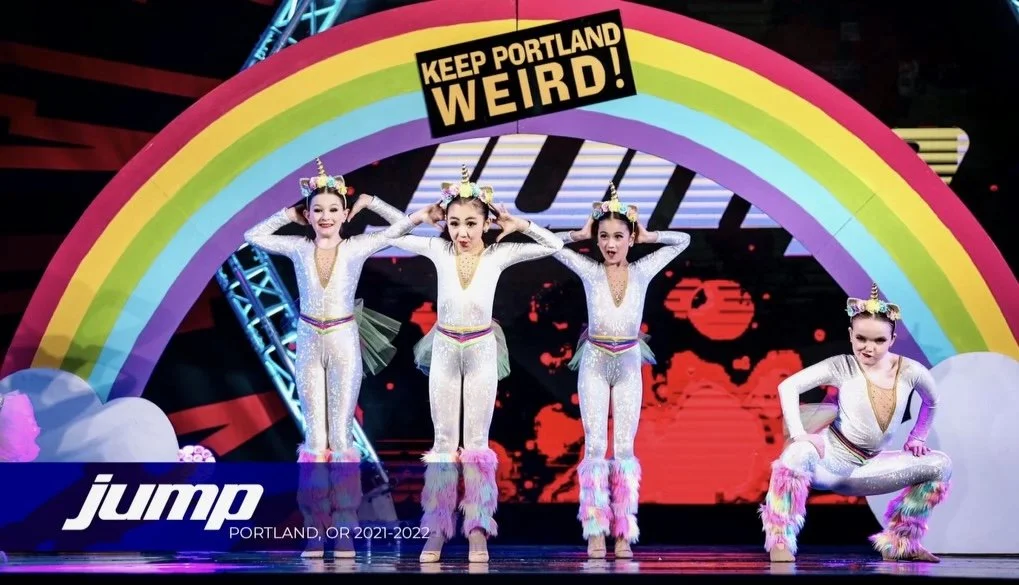 Four young girls in matching unicorn-themed costumes perform a dance on stage under a rainbow arch with a sign that reads "Keep Portland Weird."