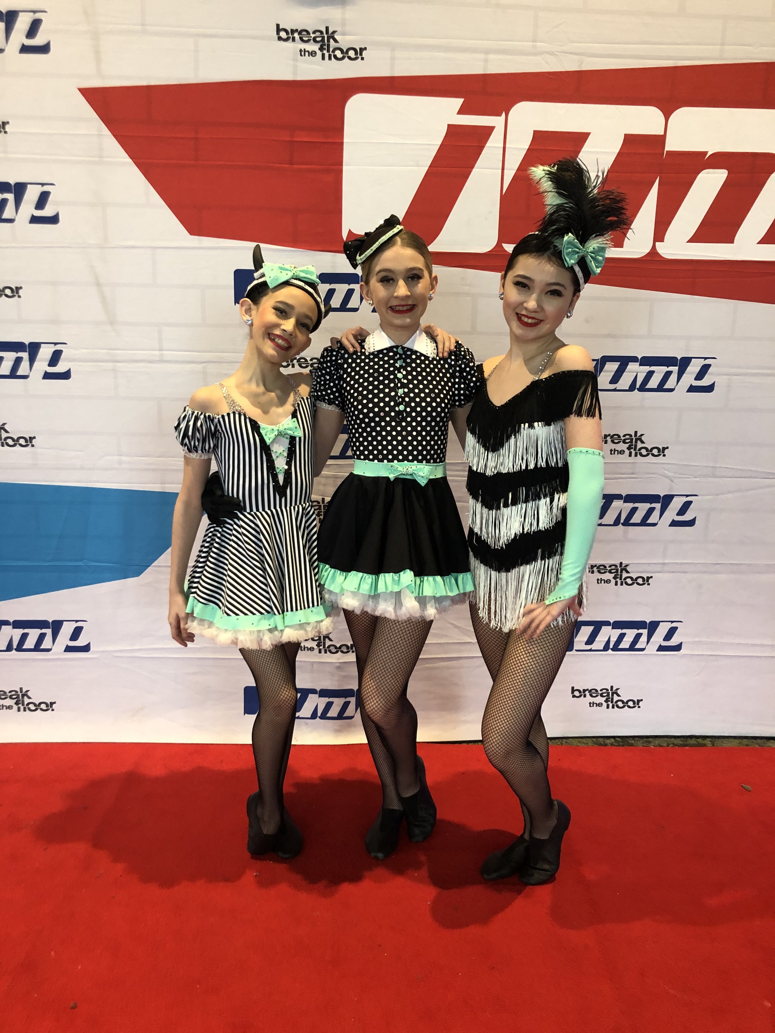 Three women dressed in vintage-style showgirl costumes with black and white striped patterns, neon green accents, and fringe, posing on a red carpet in front of a backdrop with the words 'break the floor' and 'TMC' logo.