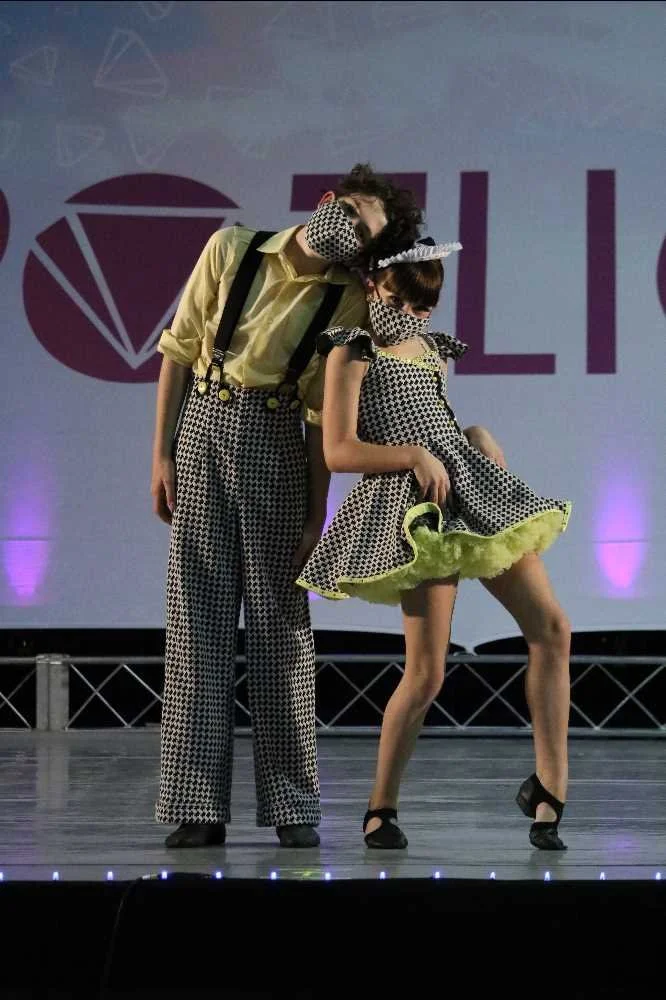Two performers on stage dressed in matching black and white patterned outfits with yellow accents, wearing masks, with one leaning on the other.