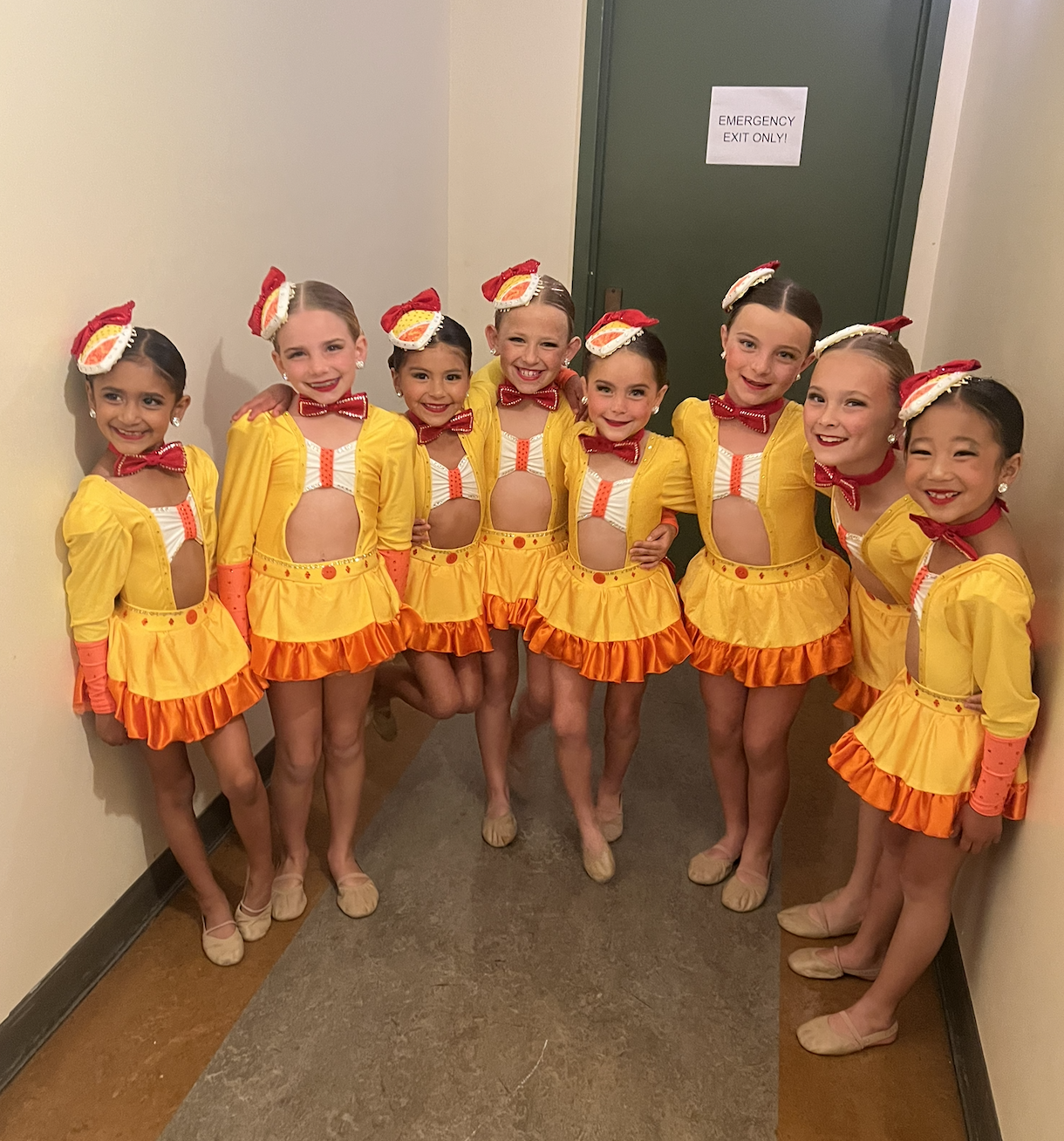 Group of young girls in bright yellow and orange dance costumes, standing side by side in a hallway, smiling and posing for the camera.