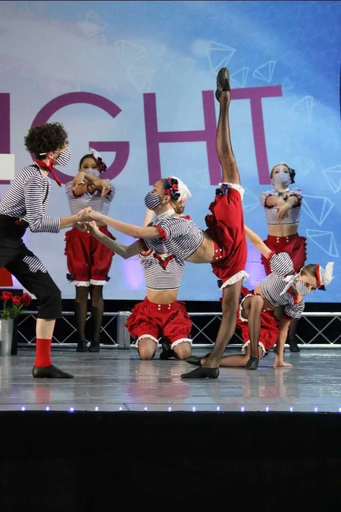 Children dressed in red and striped costumes performing a dance routine on stage, with some holding hands and others balancing or kneeling, in front of a large screen with the partially visible word 'LIGHT'.