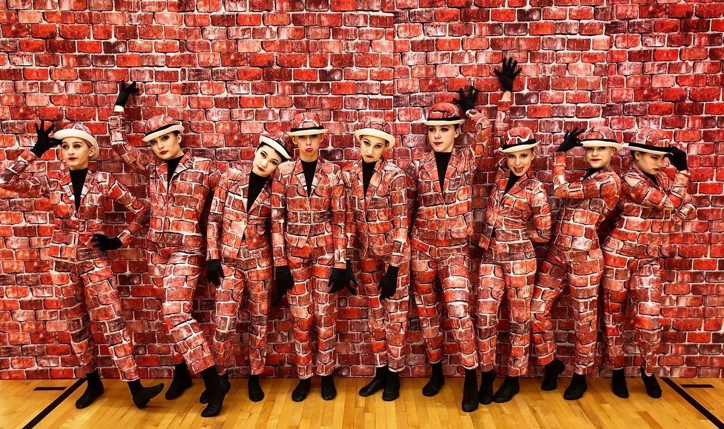 Nine women in matching 'gangster' themed costumes with brick wall background