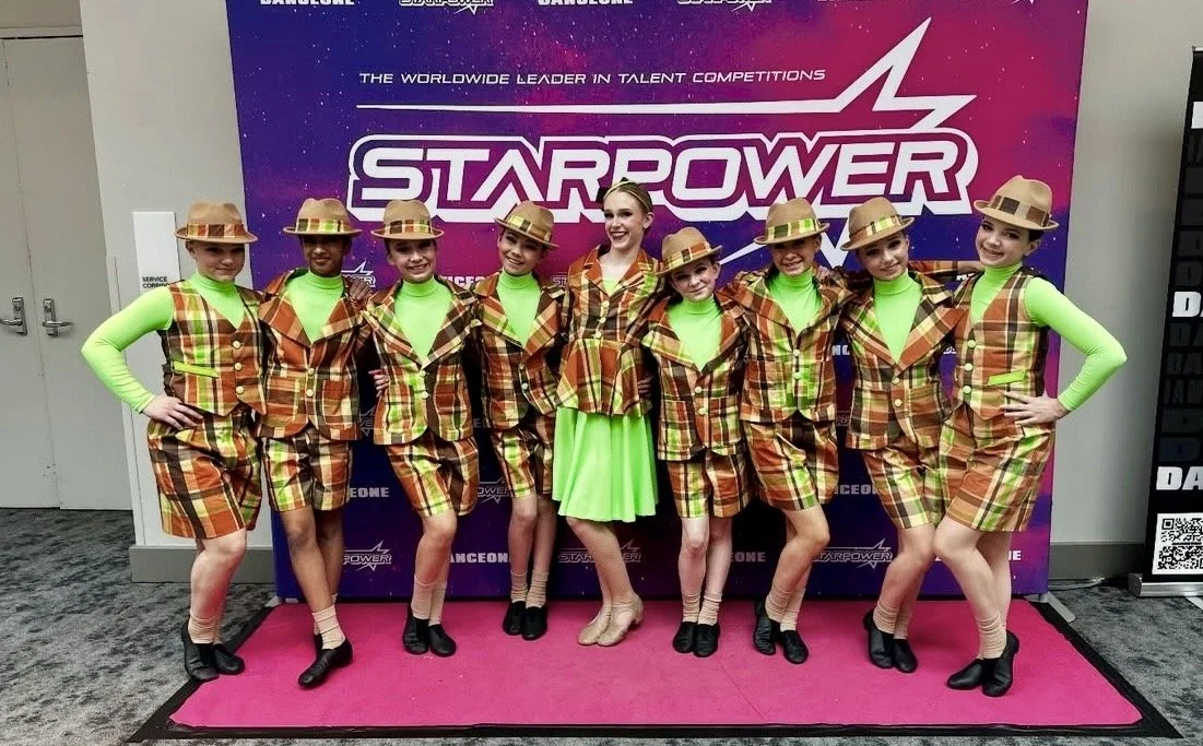 A group of young girls dressed in colorful plaid costumes with matching hats, standing in front of a Starpower competition backdrop.