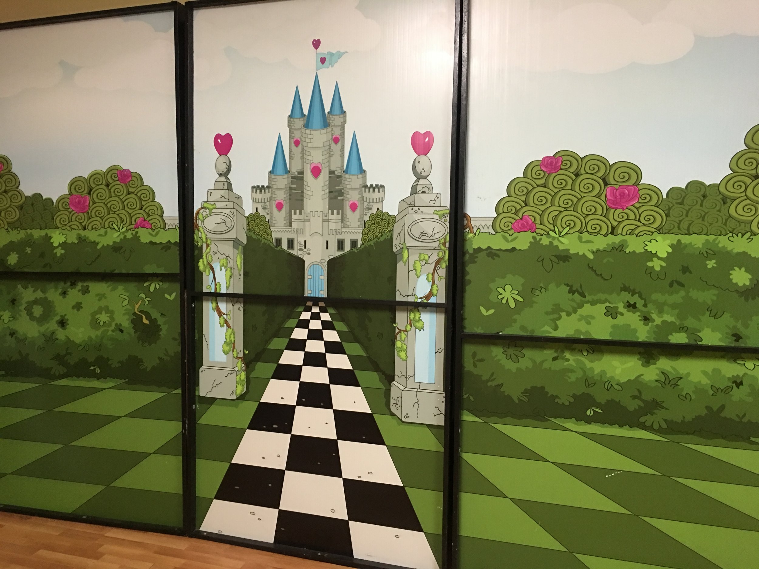 Colorful castle mural with blue towers, pink hearts, and swirling green bushes, featuring a black and white checkered path leading to the castle entrance.