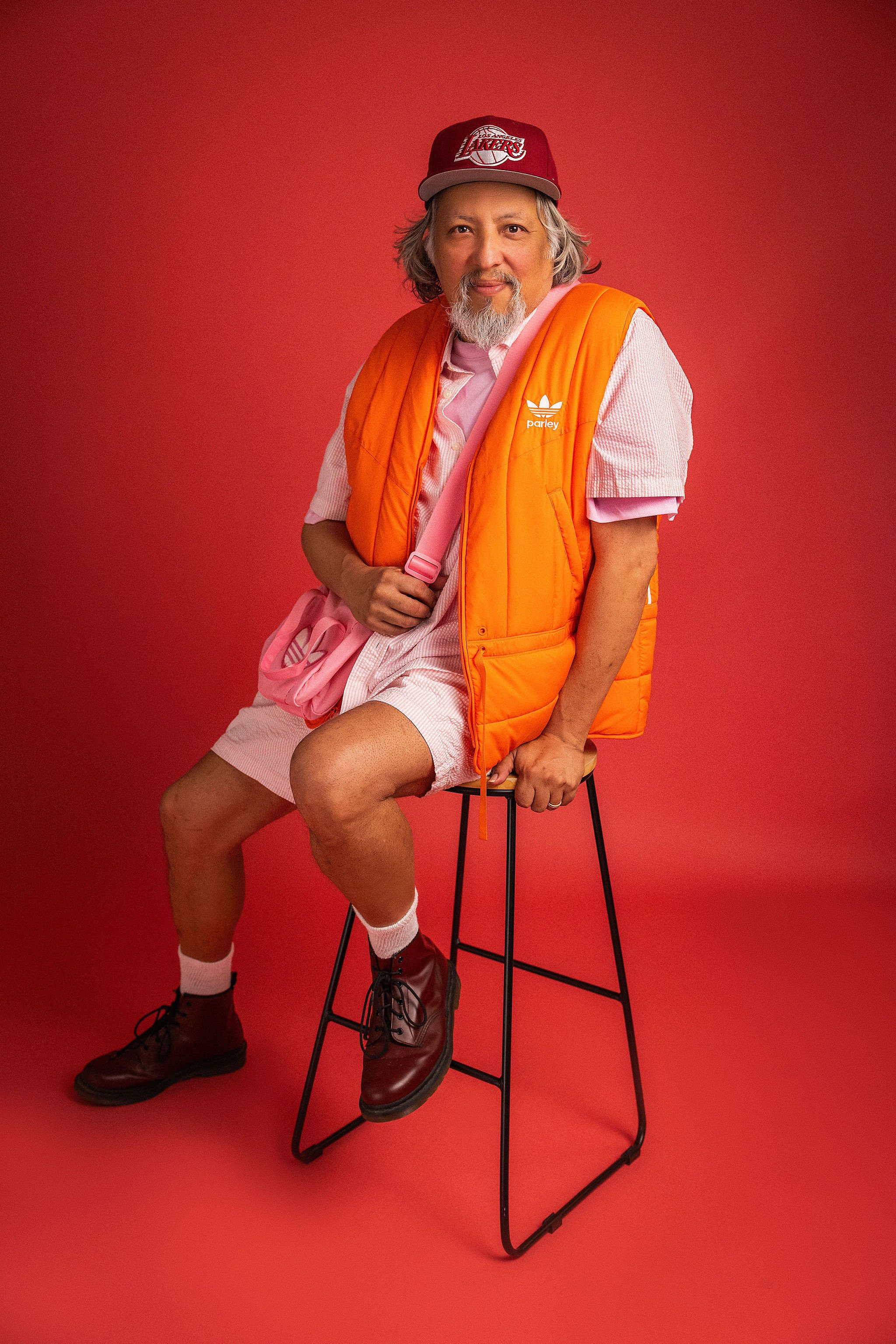 A man with gray hair, a beard, and a mustache sitting on a black stool against a red background. He is wearing a red baseball cap with a Los Angeles Lakers logo, a pink and white striped short-sleeve shirt, an orange quilted vest with the Adidas logo and the word 'parley' on it, pink shorts, white socks, and black boots. He has a pink bag with white stripes over his shoulder and is smiling subtly.