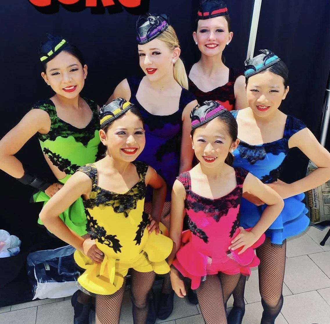 Six young girls dressed in colorful costumes and fascinators, posing in front of a black backdrop with red text.