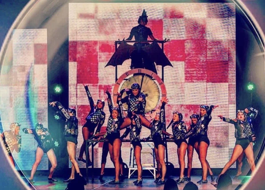 A group of female dancers perform on stage, dressed in black and white costumes with matching accessories, with a large clock and a figure holding an umbrella in the background.