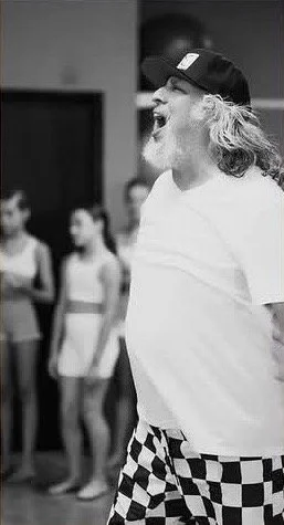 Man with long hair, beard, wearing a backwards baseball cap and checkered shorts, appears to be singing or speaking passionately, with a group of young girls standing in the background.