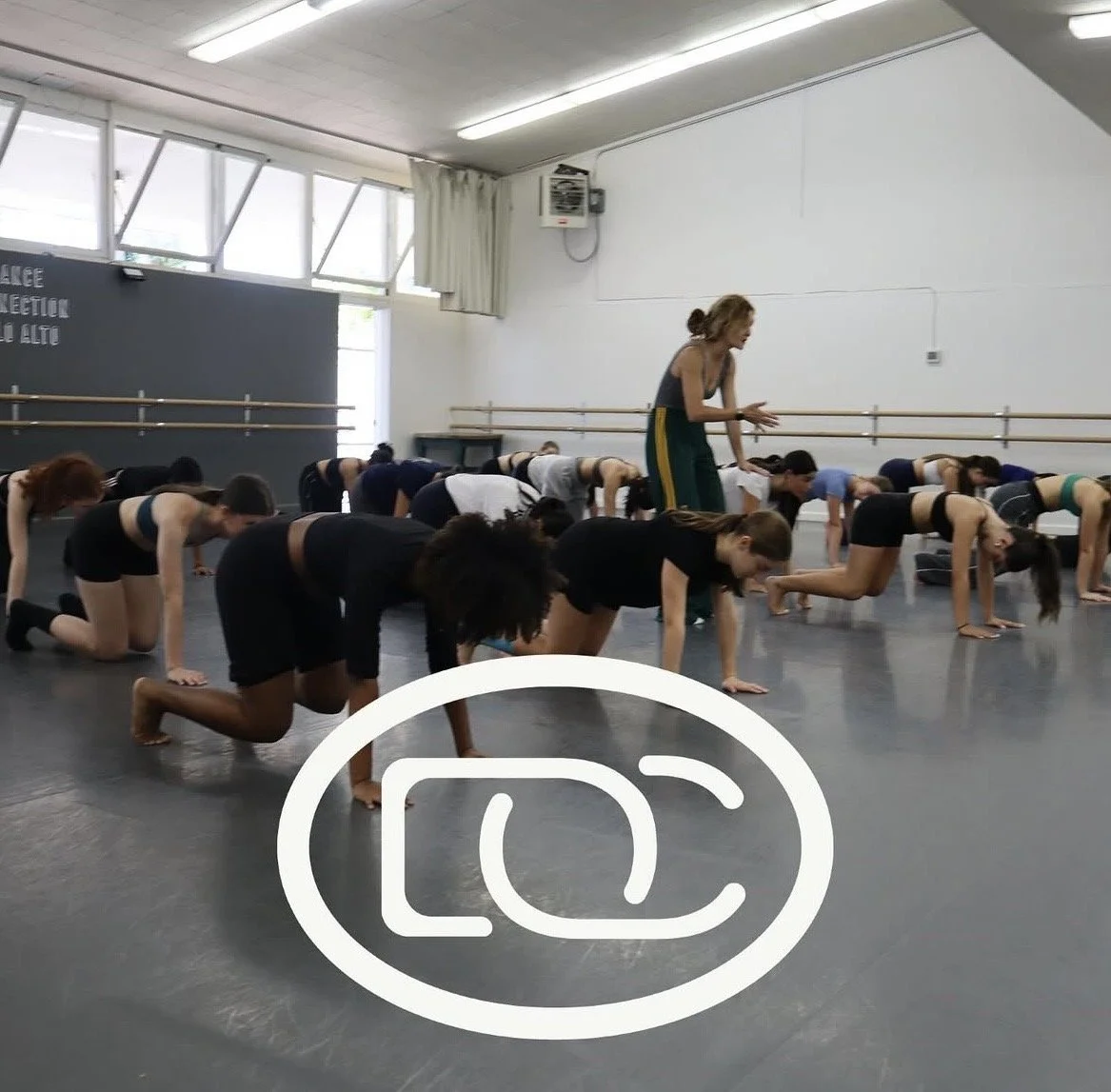 A group of dancers practicing in a studio with large windows, bending forward on all fours, as an instructor guides them.