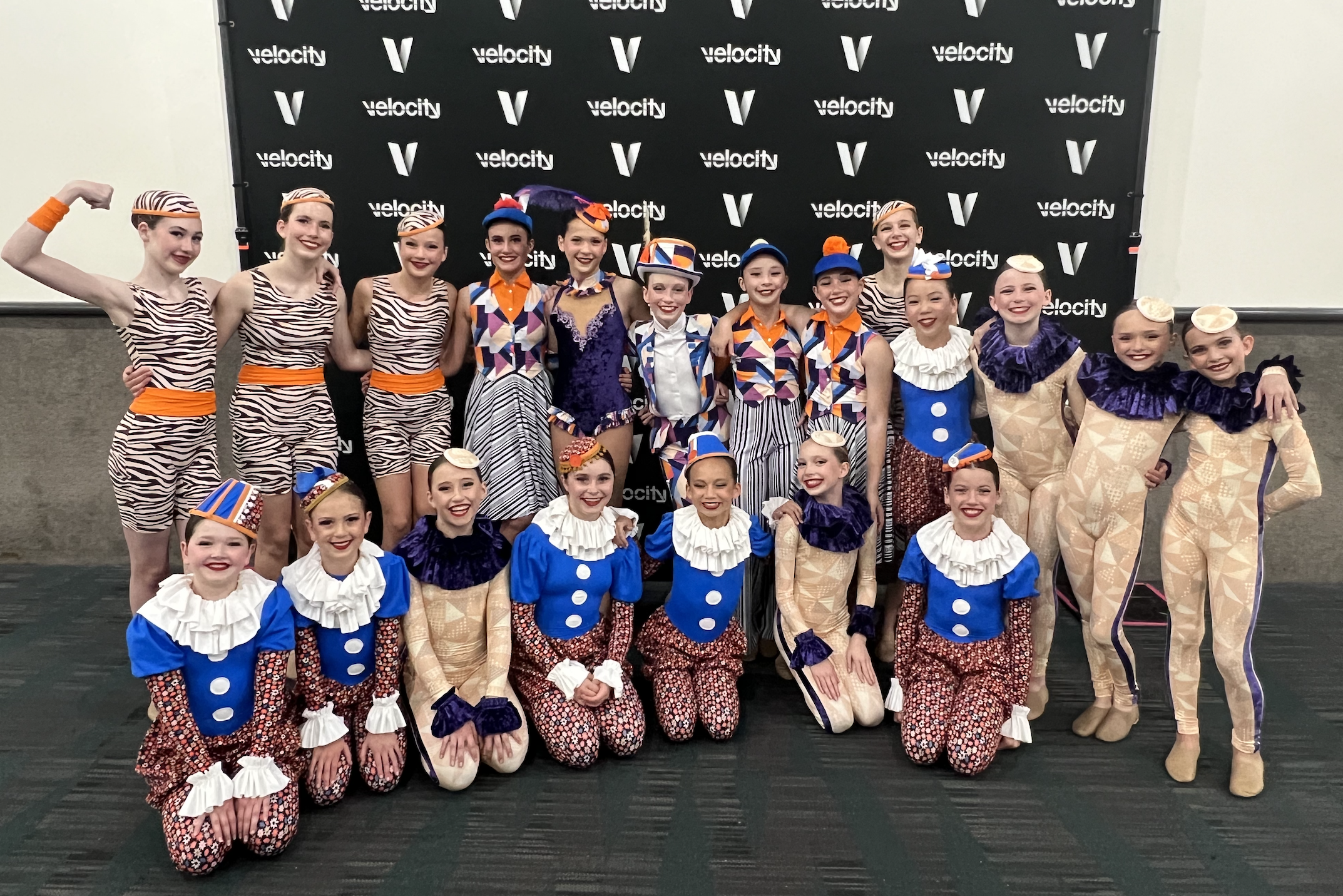 A group of young dancers dressed in colorful costumes, some with zebra-stripe outfits and others with patterned suits, posing together in front of a black backdrop with the 'Velocity' logo, smiling at a dance competition or performance event.
