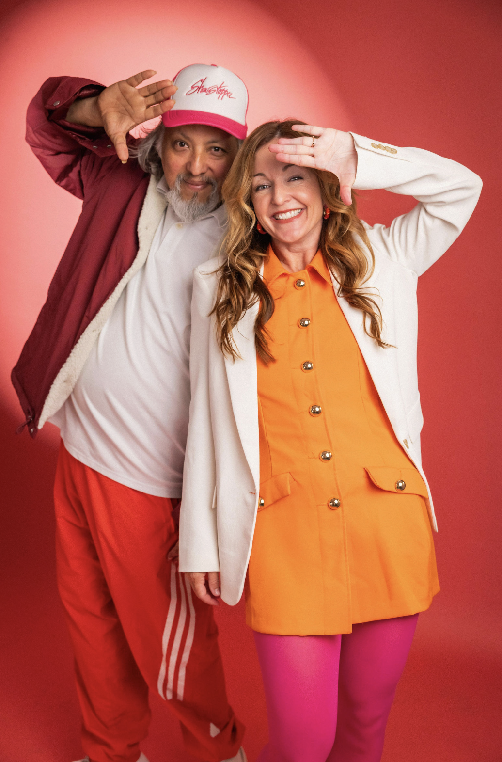 A man and a woman pose together with the man saluting and the woman smiling, both dressed in colorful retro-inspired outfits against a pink background.