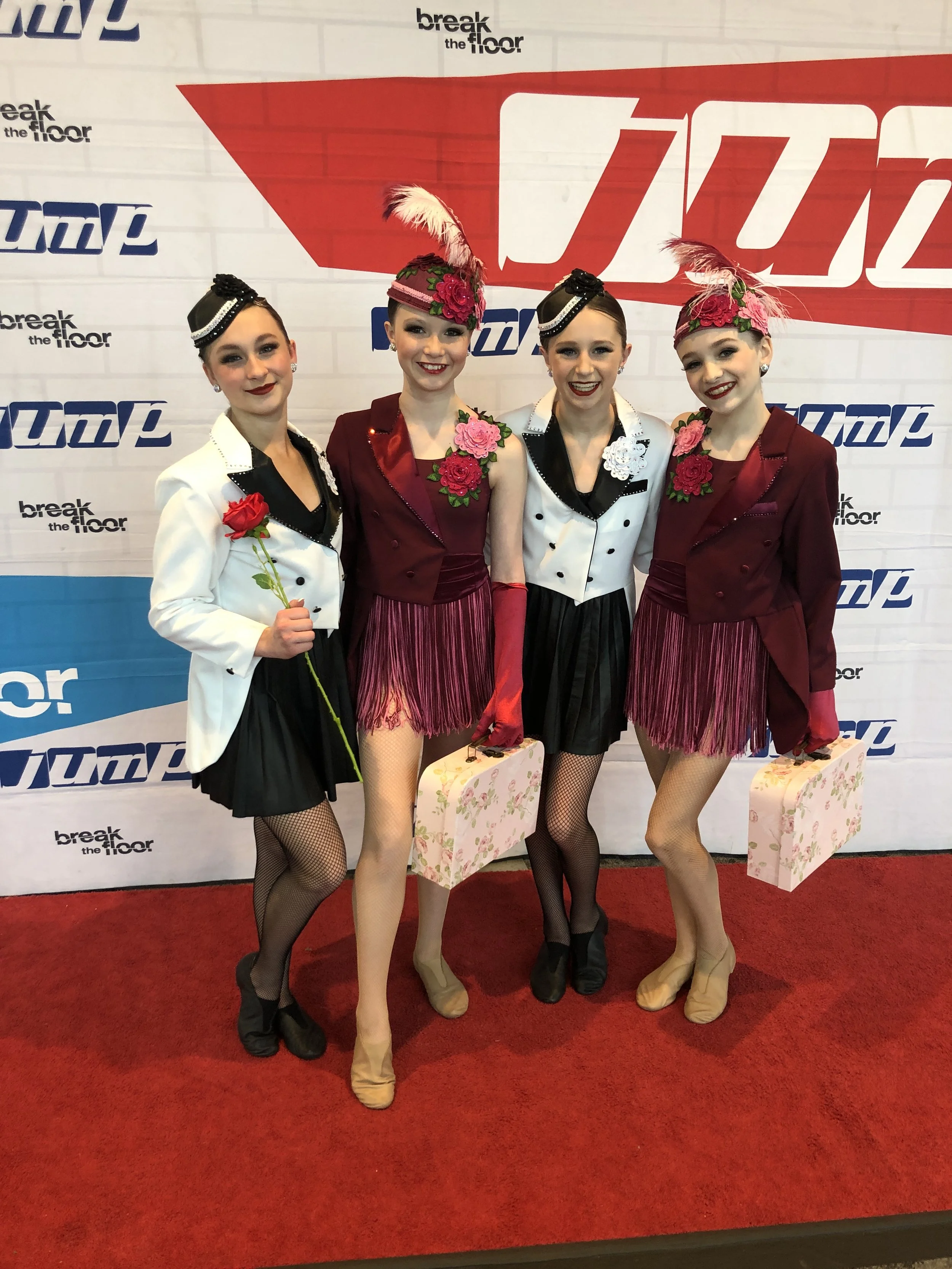 Four young women dressed in vintage-style costumes, standing on a red carpet in front of a backdrop with logos that say 'break the floor' and 'JUMP'. They are smiling, holding floral suitcases, and some are holding red roses, with one wearing a color
