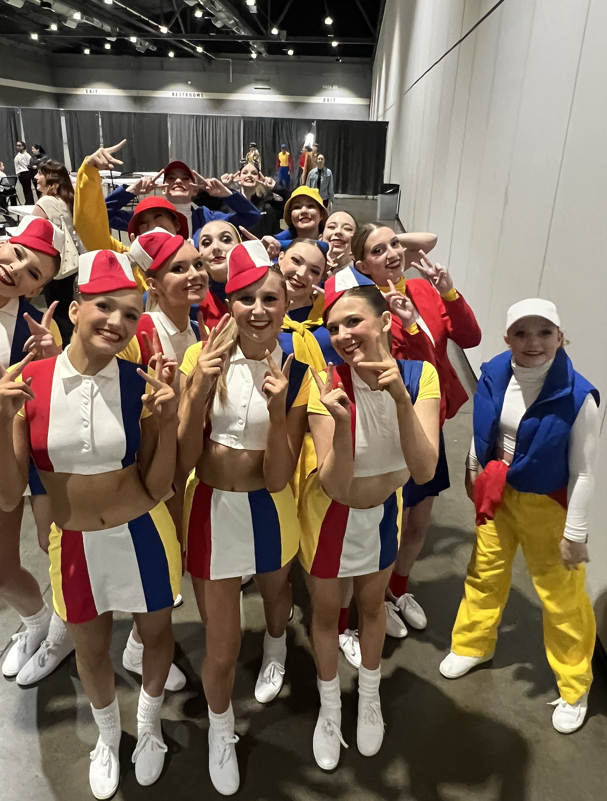 Group of dancers in colorful costumes posing backstage, smiling and making peace signs.