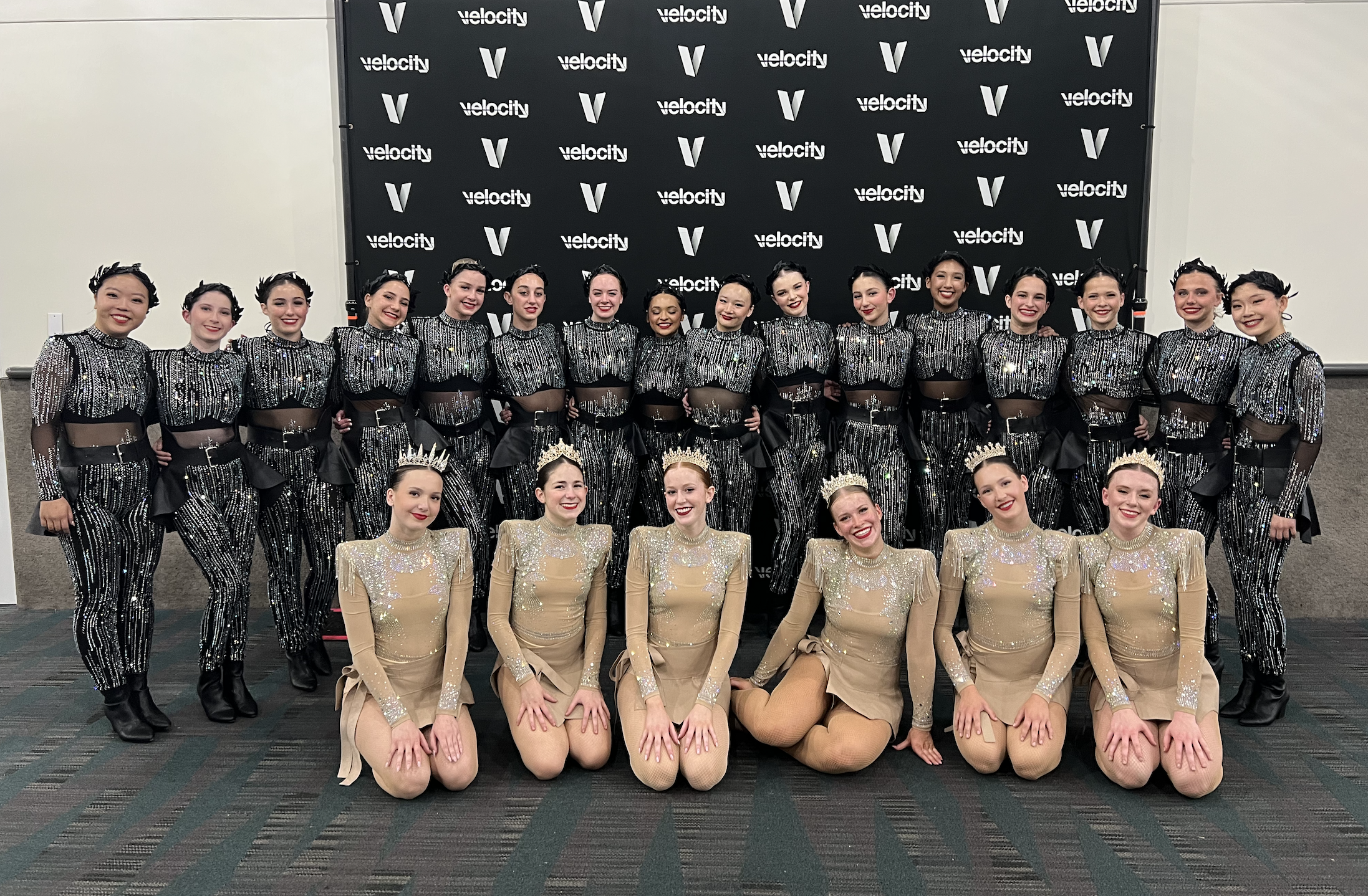 Group of young women in dance costumes posing together indoors with a black backdrop displaying the logos 'velocity' and a V shape