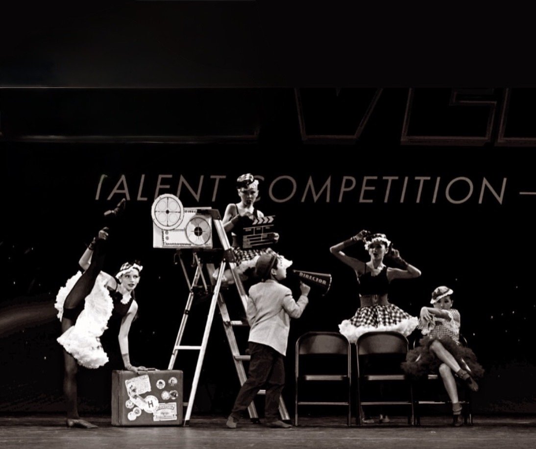 Black and white photo of children performing on stage, dressed in vintage costumes, with some sitting on or standing near chairs, and one child holding a clapperboard, during a talent competition.
