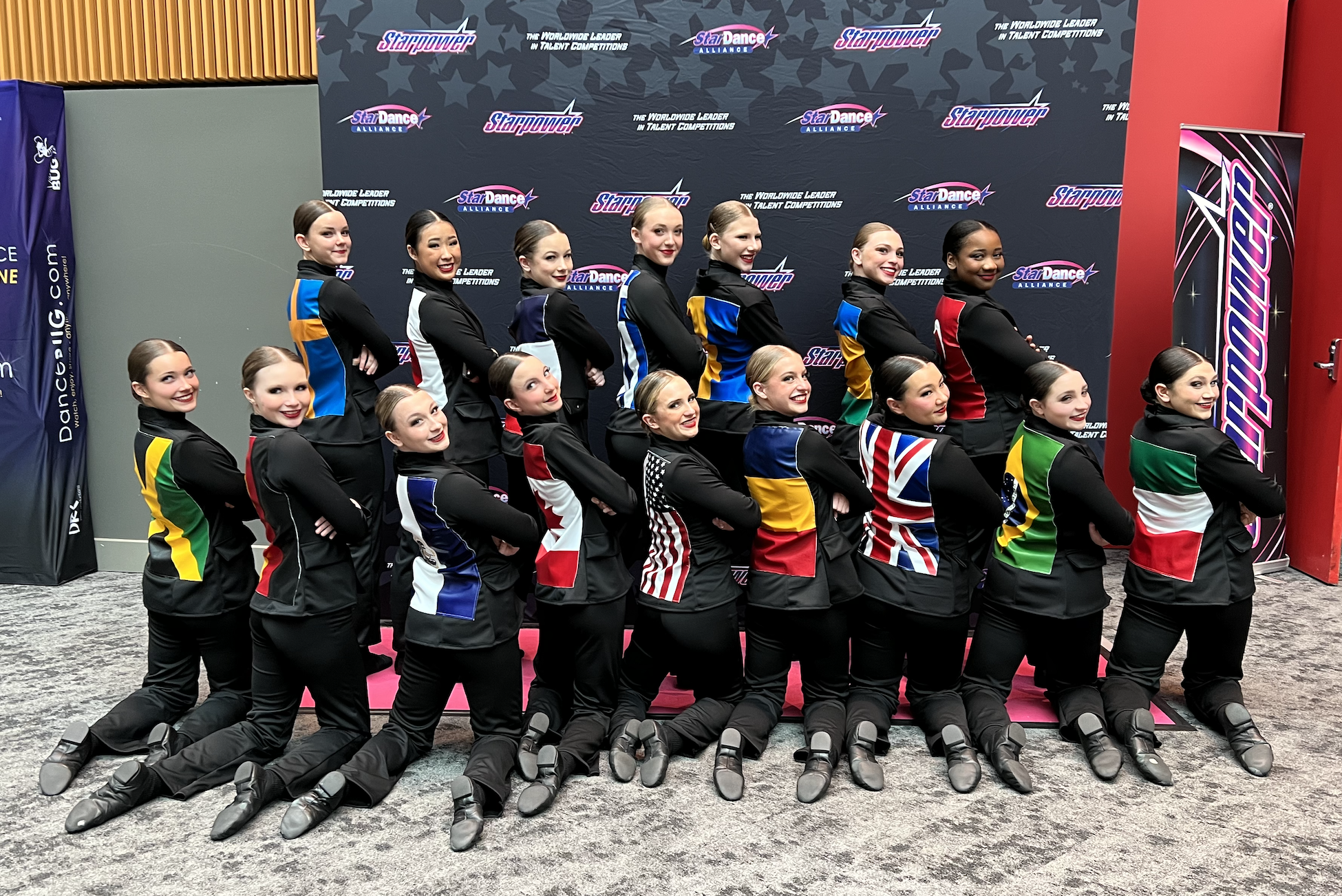 A group of female dancers in colorful costumes with flags, posing in front of a backdrop with logos for Starpower and StarDance Alliance, at a dance competition.