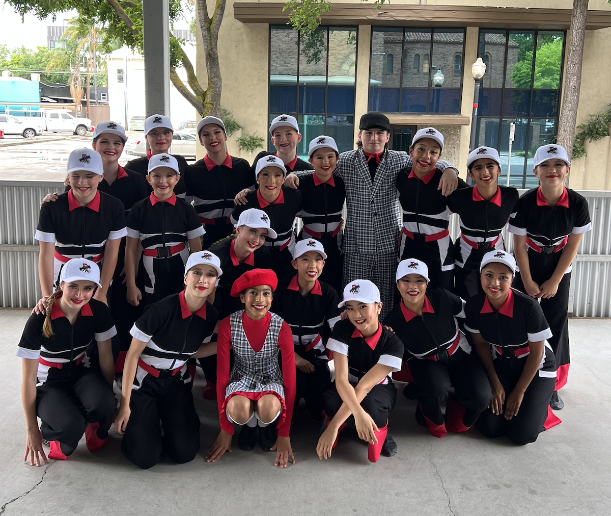 Group of young dancers in black, red, and white costumes with caps, posing outdoors with a woman in a checkered outfit. Some are kneeling, others standing, smiling for the camera.