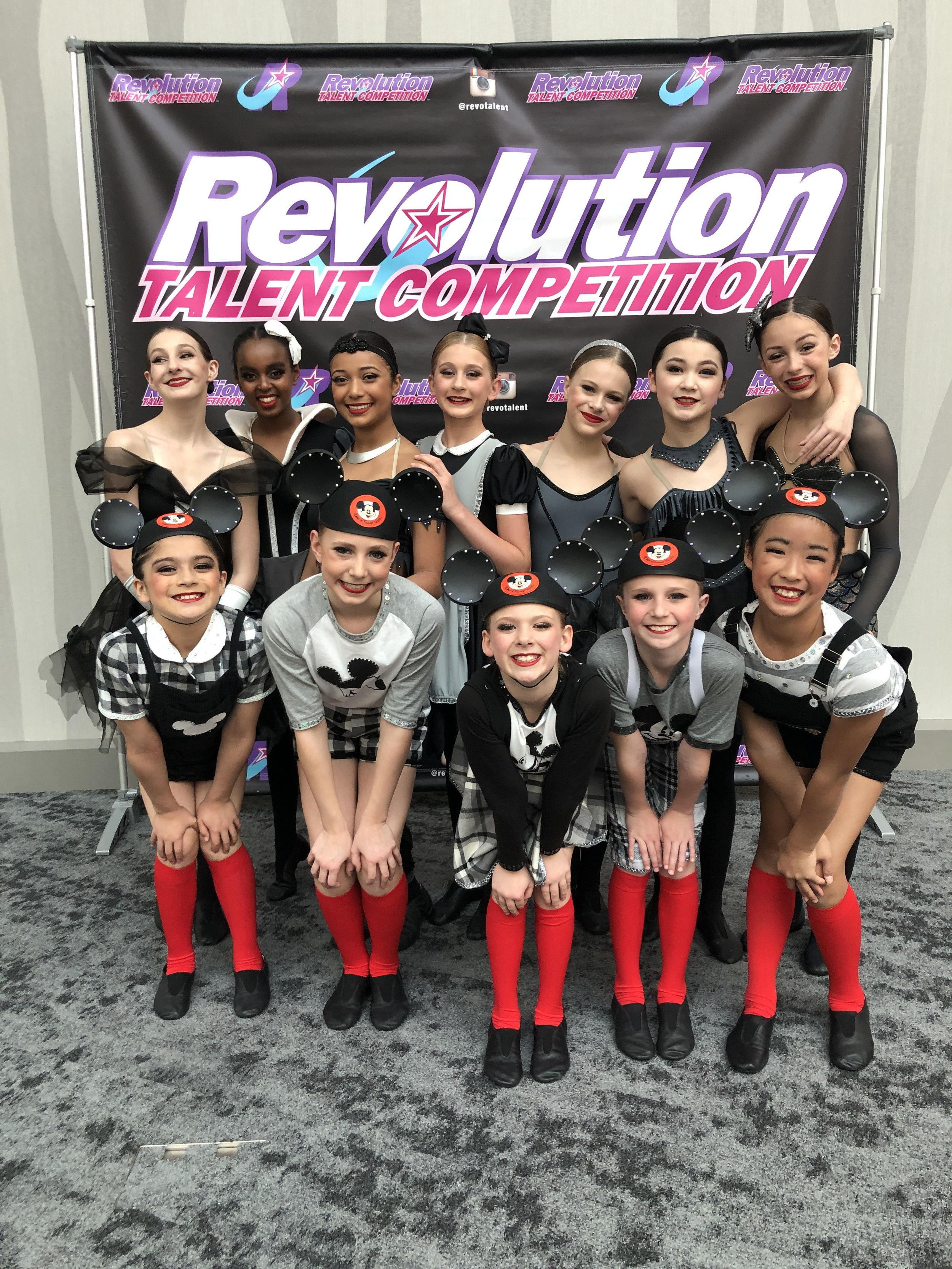 Group of young girls in dance costumes posing in front of a "Revolution Talent Competition" banner at a dance event.