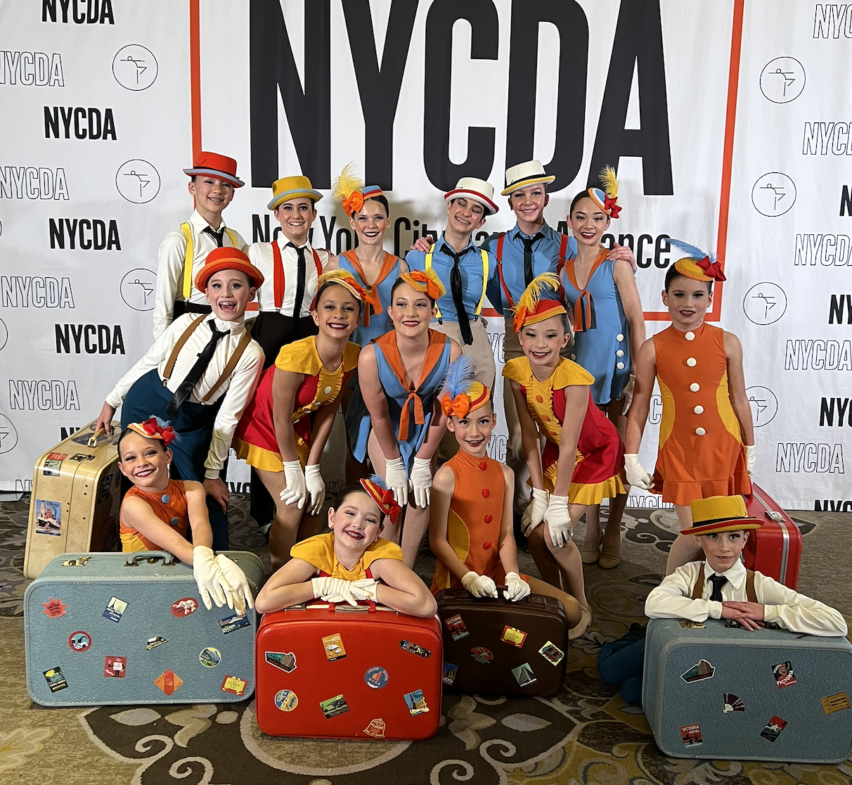 Group of young girls in colorful costumes with hats and gloves at NYCDA event, holding vintage suitcases, standing in front of NYCD logo backdrop.