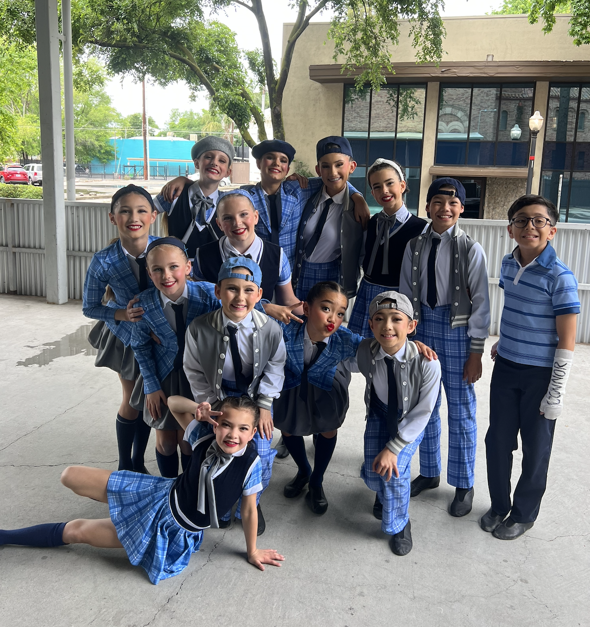A group of children dressed in blue and gray costumes, possibly for a play or dance, gathered outdoors on a concrete surface with a building, trees, and a street in the background.