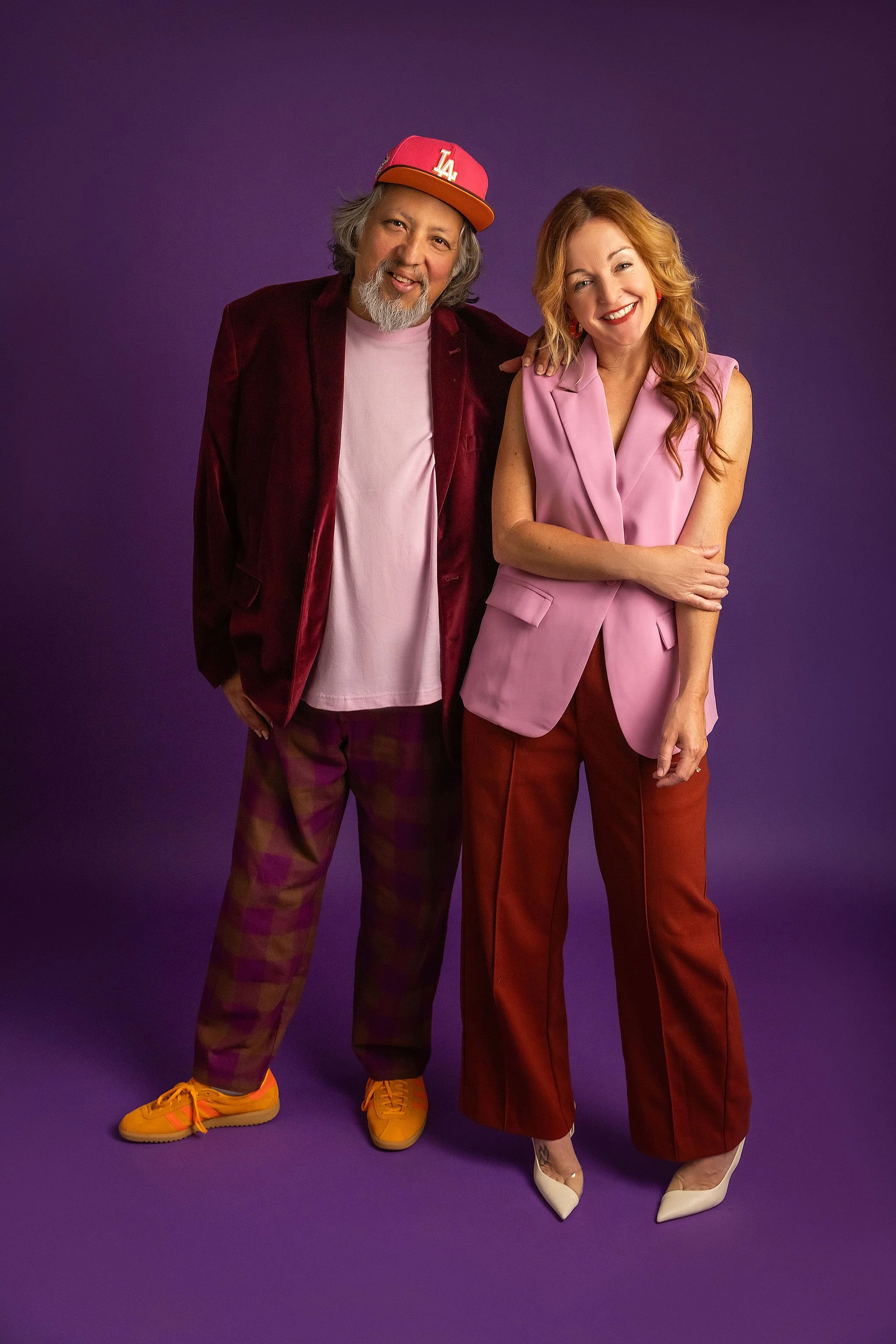 Two people standing together against a purple background, smiling. The person on the left is wearing a red LA baseball cap, burgundy jacket, plaid pants, and yellow sneakers. The person on the right is wearing a pink sleeveless blazer, red pants, and beige high heels.