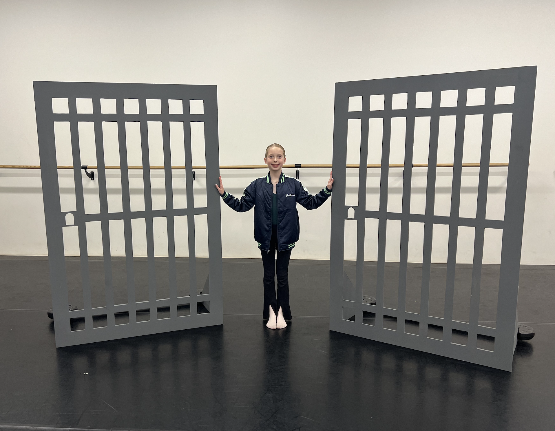 A young dancer standing between two large gray prison cell cutouts, smiling, in a dance studio with a white wall and a wooden ballet barre.