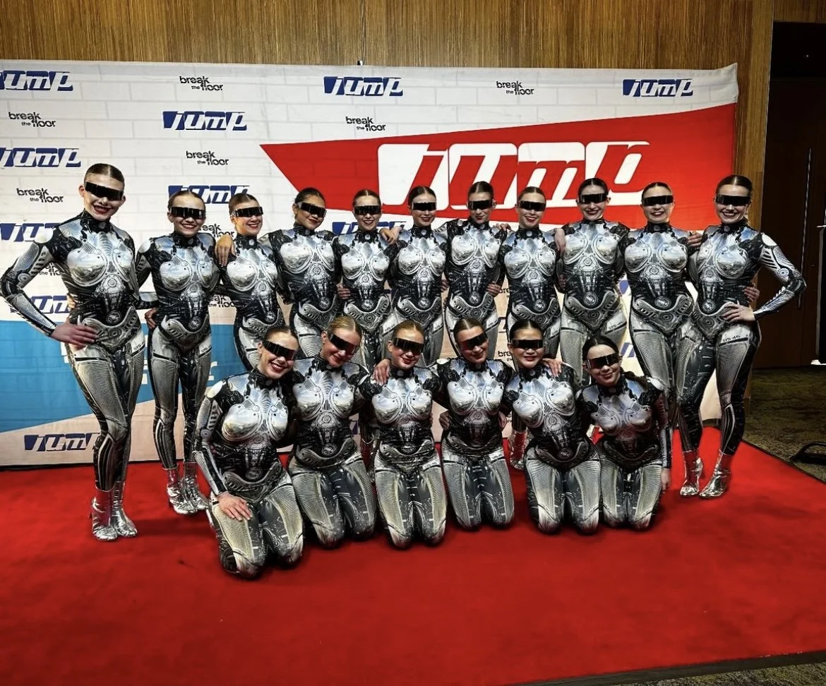Group of 15 women dressed in silver and black futuristic costumes, wearing sunglasses, posing on a red carpet in front of a backdrop with logos.