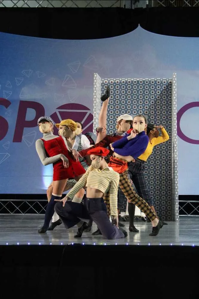 Group of six young women performing a dance on stage, dressed in colorful, vintage-inspired outfits, with a patterned backdrop and stage lights.