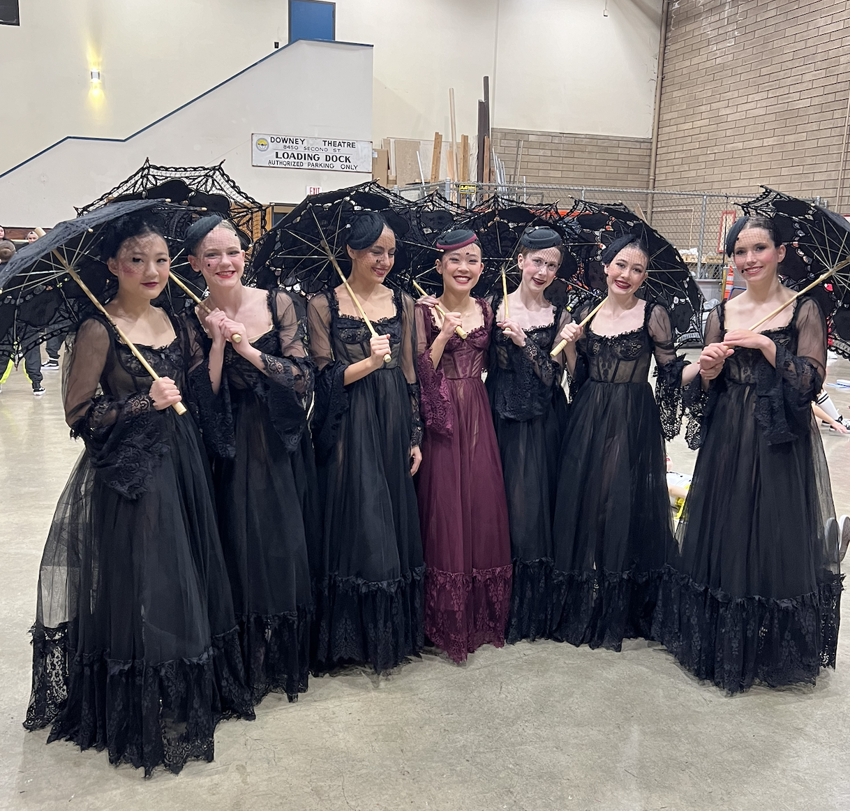 Seven women dressed in black and burgundy vintage-style dresses, holding black lace umbrellas, standing in an indoor event space.