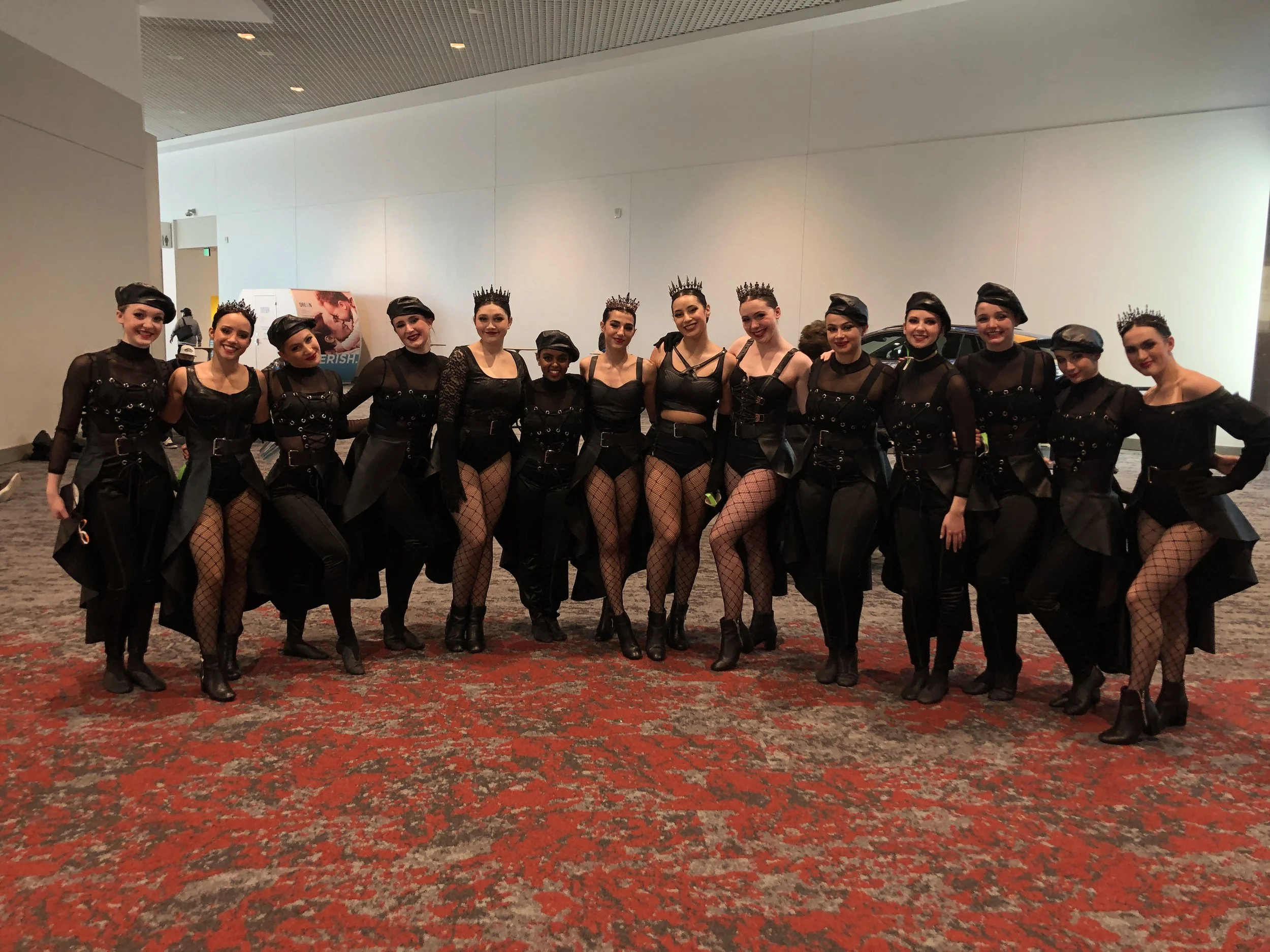 A group of fifteen women dressed in black costumes with fishnet stockings, some with crowns, standing together indoors on a red patterned carpet, smiling at the camera.