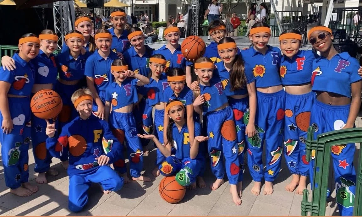 Group of young girls dressed in blue dance costumes with colorful patches, wearing orange headbands, posing together outdoors during daytime with some holding basketballs.