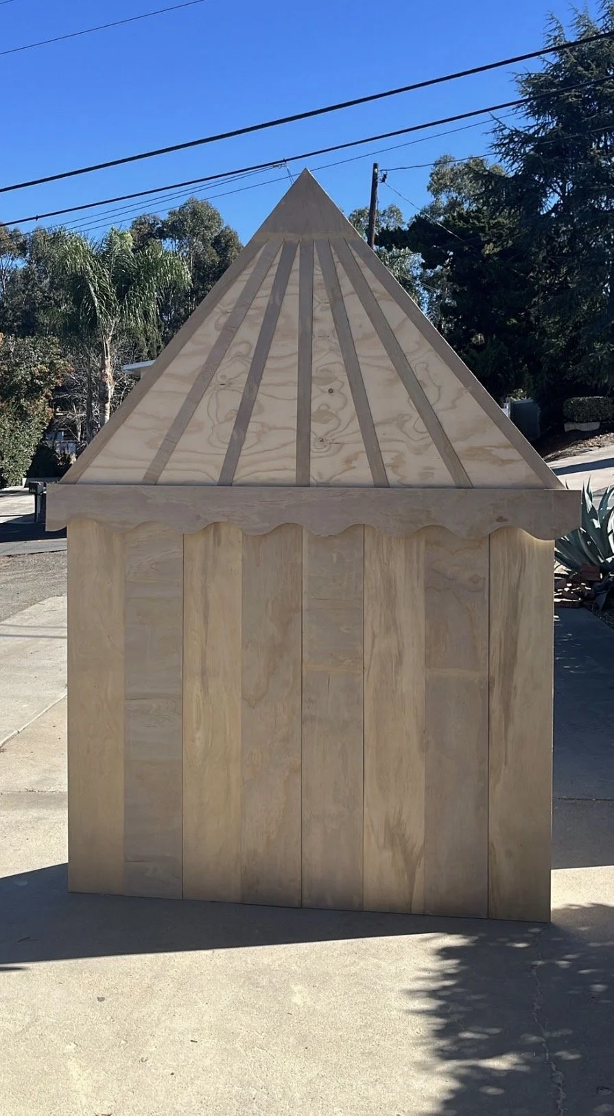 Unfinished wooden playhouse or shed with a peaked roof, installed outdoors on a concrete surface, surrounded by trees and a clear blue sky.