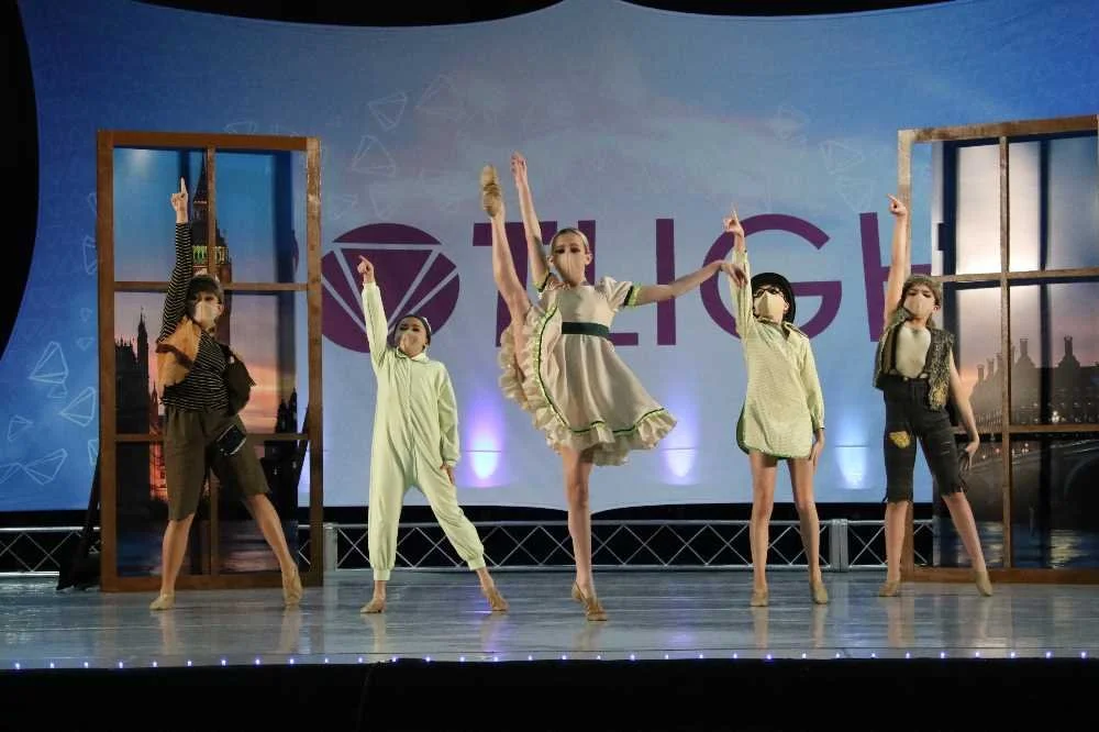Five young girls in dance costumes performing on stage with a backdrop that reads "LIGHT" and cityscape images, some dancers raising their arms.