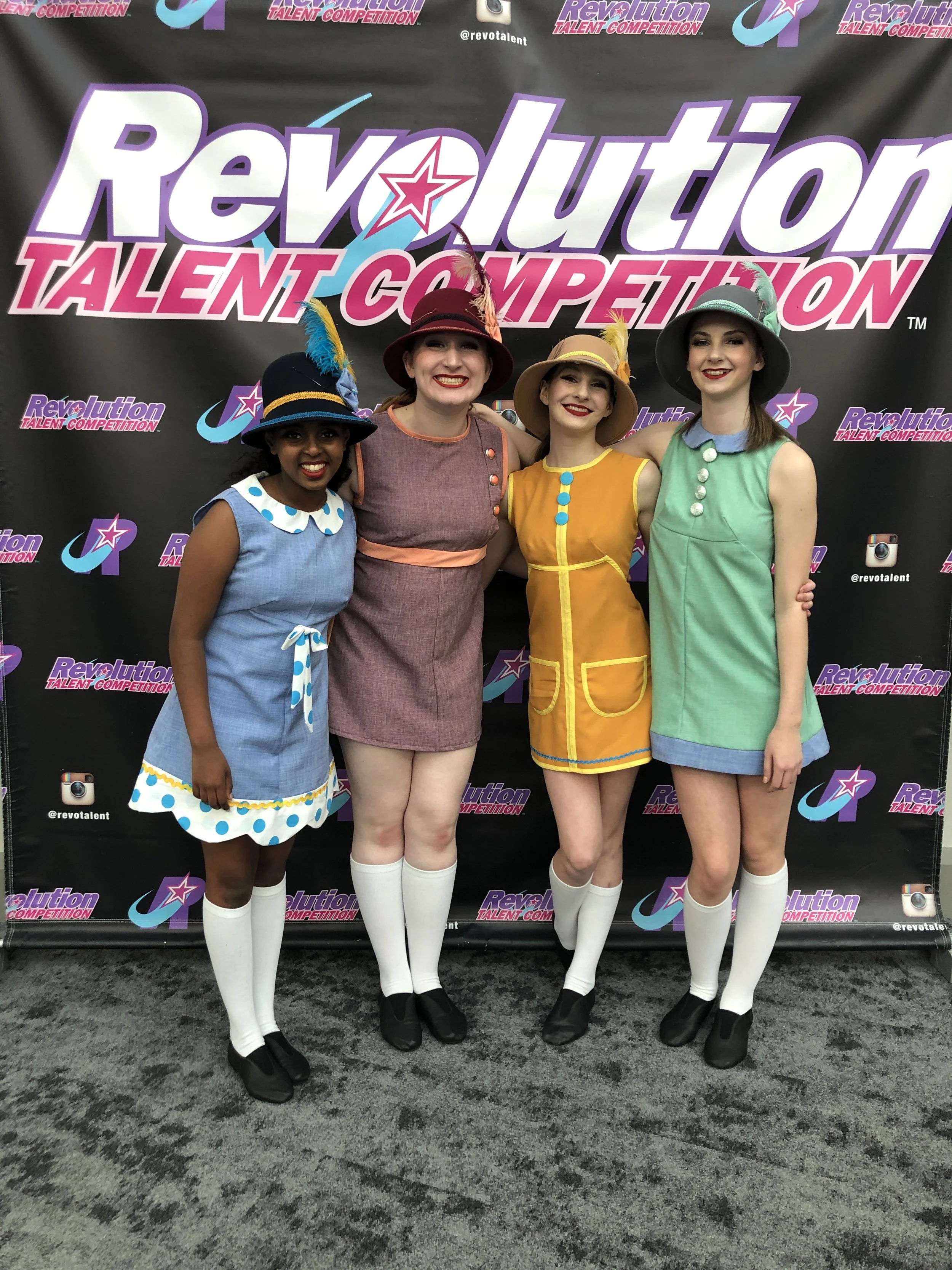 Four women dressed in colorful costumes with feathered hats, smiling and standing in front of a backdrop that says 'Revolution Talent Competition' and has social media icons.