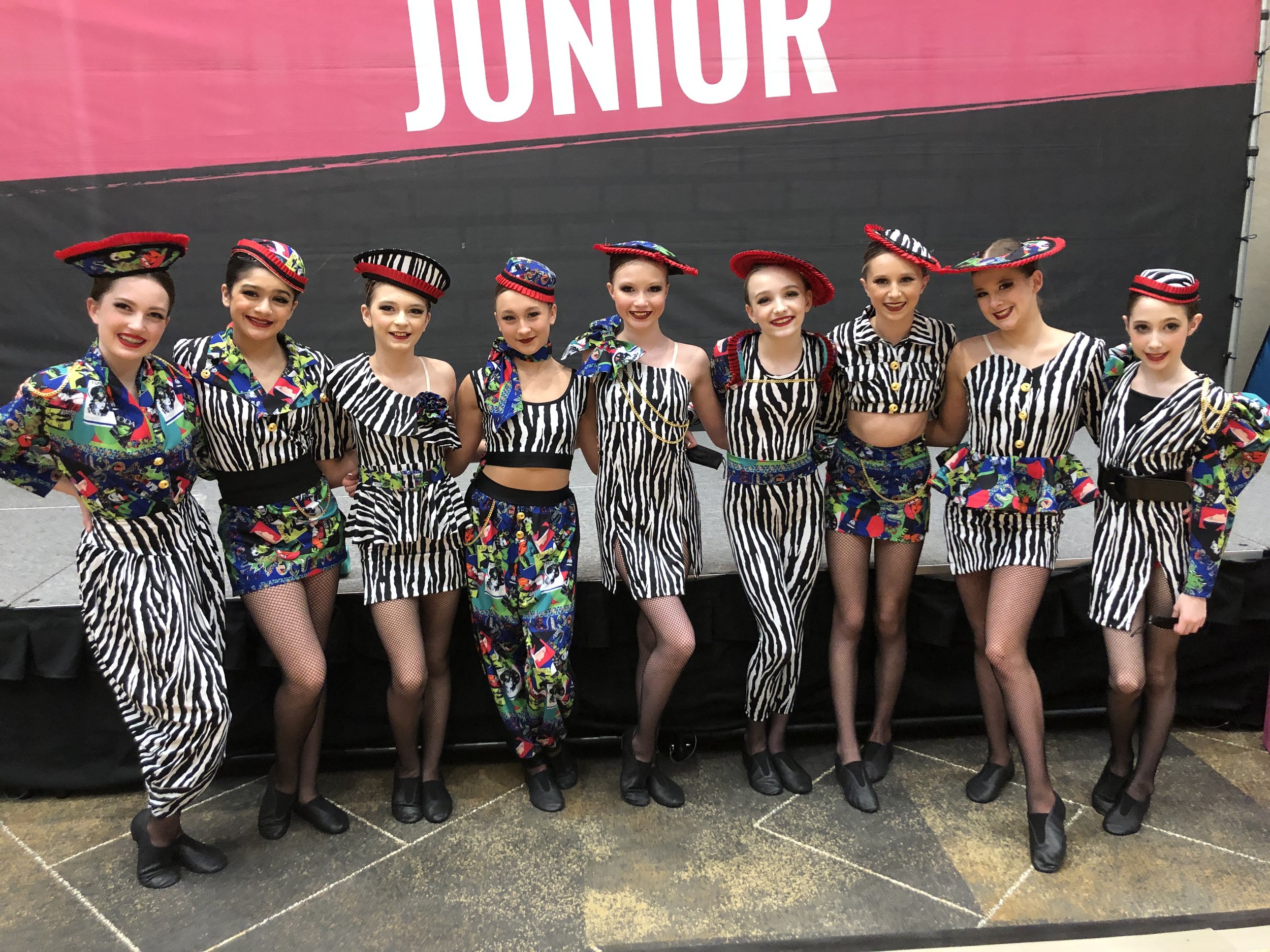 A group of ten young female dancers dressed in colorful costumes with black and white patterns, wearing matching hats, standing side by side and posing for a photo in front of a black stage backdrop with a pink sign that reads 'JUNIOR' at an event or
