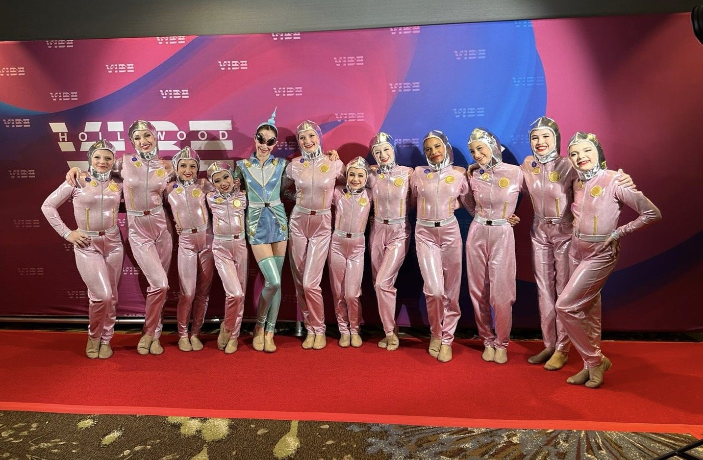 Group of eleven performers in pink and blue costumes posing on a red carpet in front of a pink and blue background with the words 'VIBE' and 'HOLLYWOOD'.