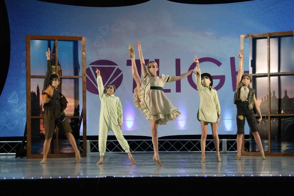 Five young girls performing a dance on stage with a city skyline backdrop.