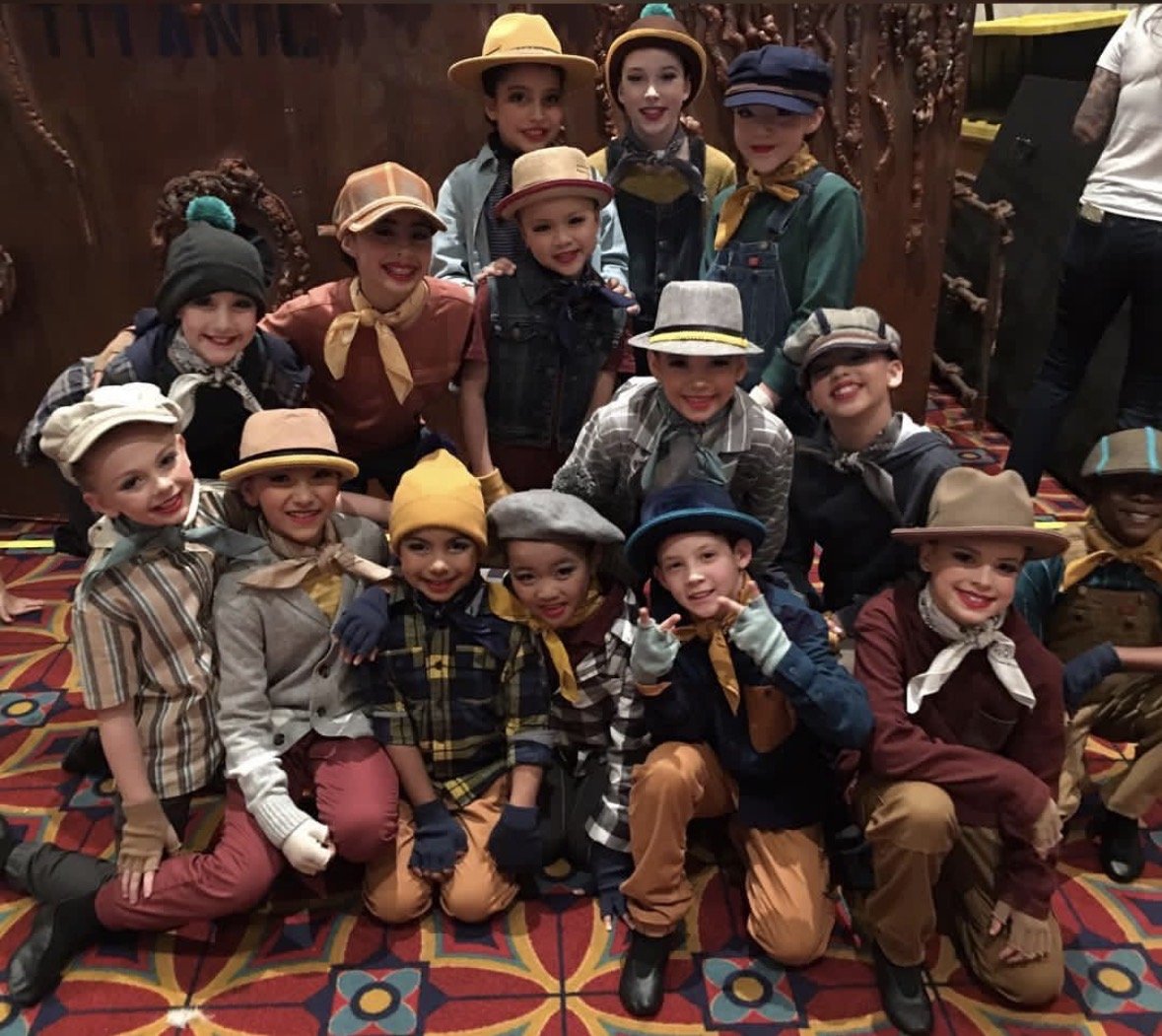Group of children dressed in vintage Western outfits with hats, bandanas, and gloves, posing together indoors with a decorative wooden wall and a colorful patterned carpet.