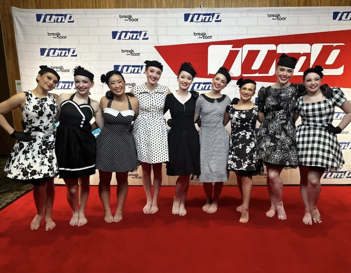 Nine women dressed in vintage 1950s style outfits, standing on a red carpet in front of a backdrop with logos. They are smiling and pose for a group photo.