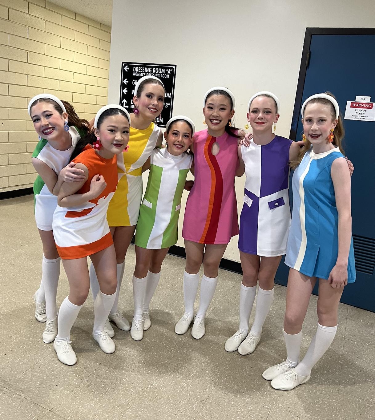 Group of young girls wearing colorful retro-style dresses with white knee-high socks and white shoes, standing in a hallway, smiling at the camera.