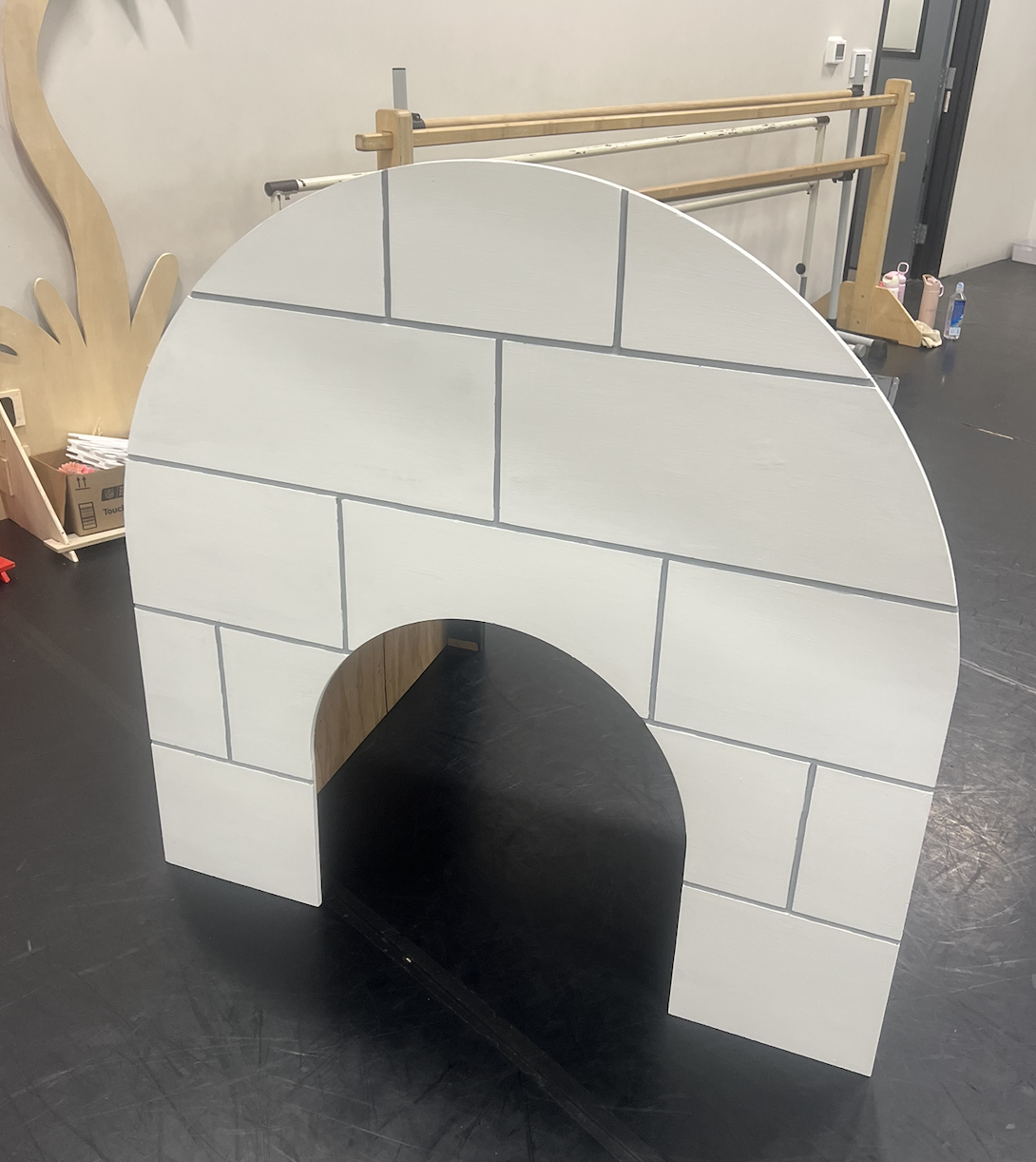 A small cardboard or wood playhouse resembling an igloo with a white brick pattern, placed on a black floor in an indoor space.