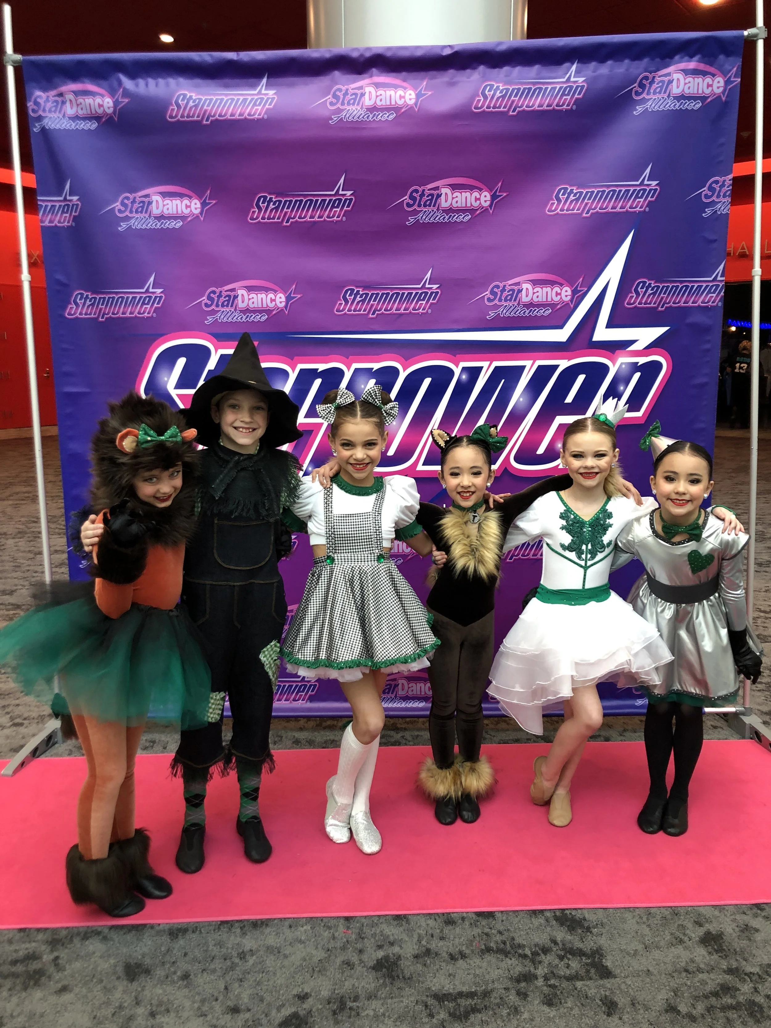 Group of five young girls dressed in colorful costumes, standing on a pink carpet in front of a promotional backdrop for Starpower StarDance Alliance, smiling and posing for the photo.