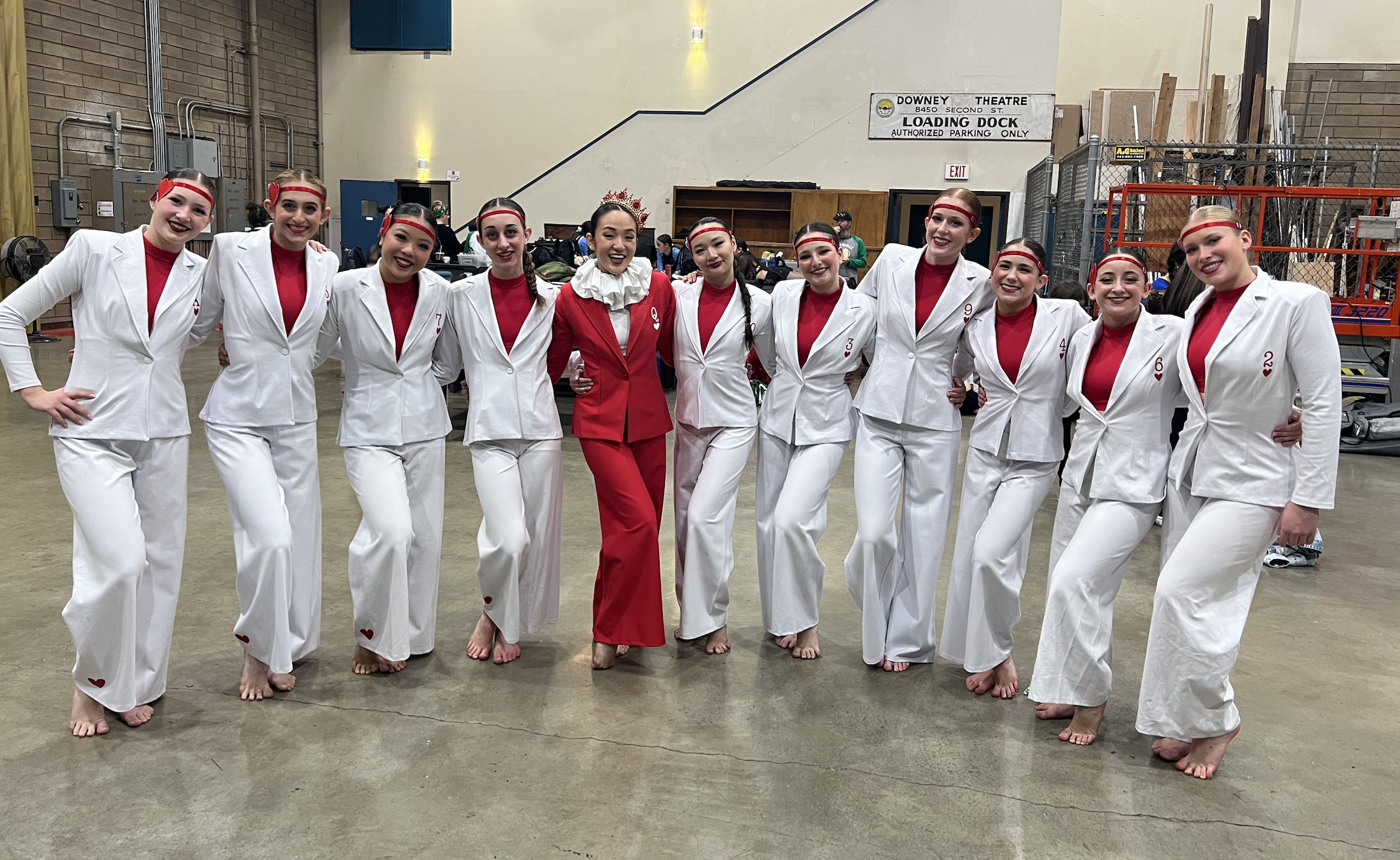A group of nine young women dressed in white suits with red shirts underneath, standing together in a line with their arms around each other's shoulders. They are smiling and appear to be backstage or in a large indoor venue, possibly a theater or pe